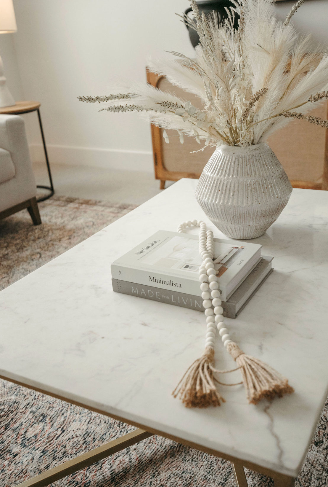 Marble coffee table styled with stacked books, beaded garland, and white textured vase with dried pampas grass in boho living room.