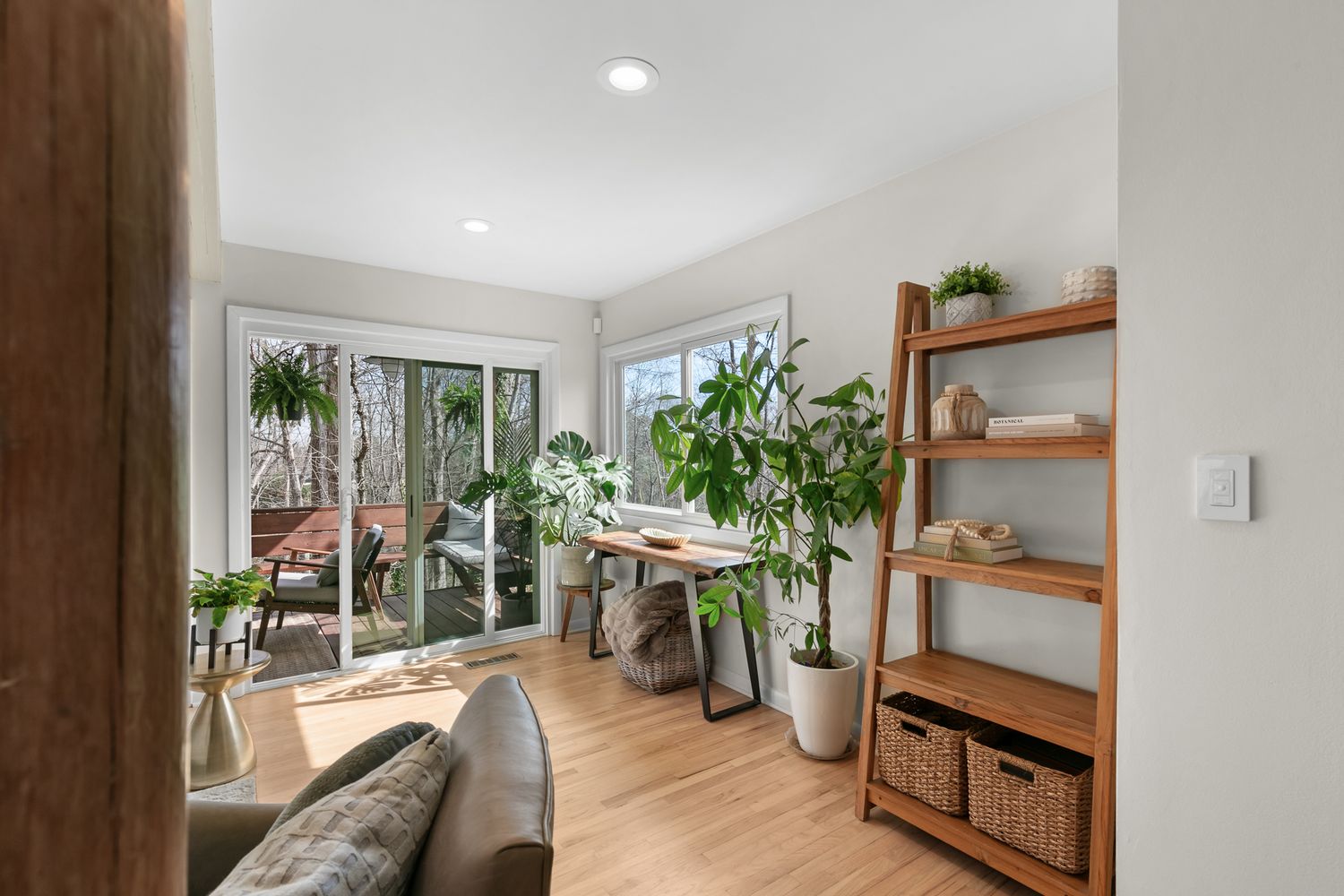 Bright home office with hardwood floors, lush indoor plants, a wooden desk, leaning shelf, and sliding glass doors to a wooded view.