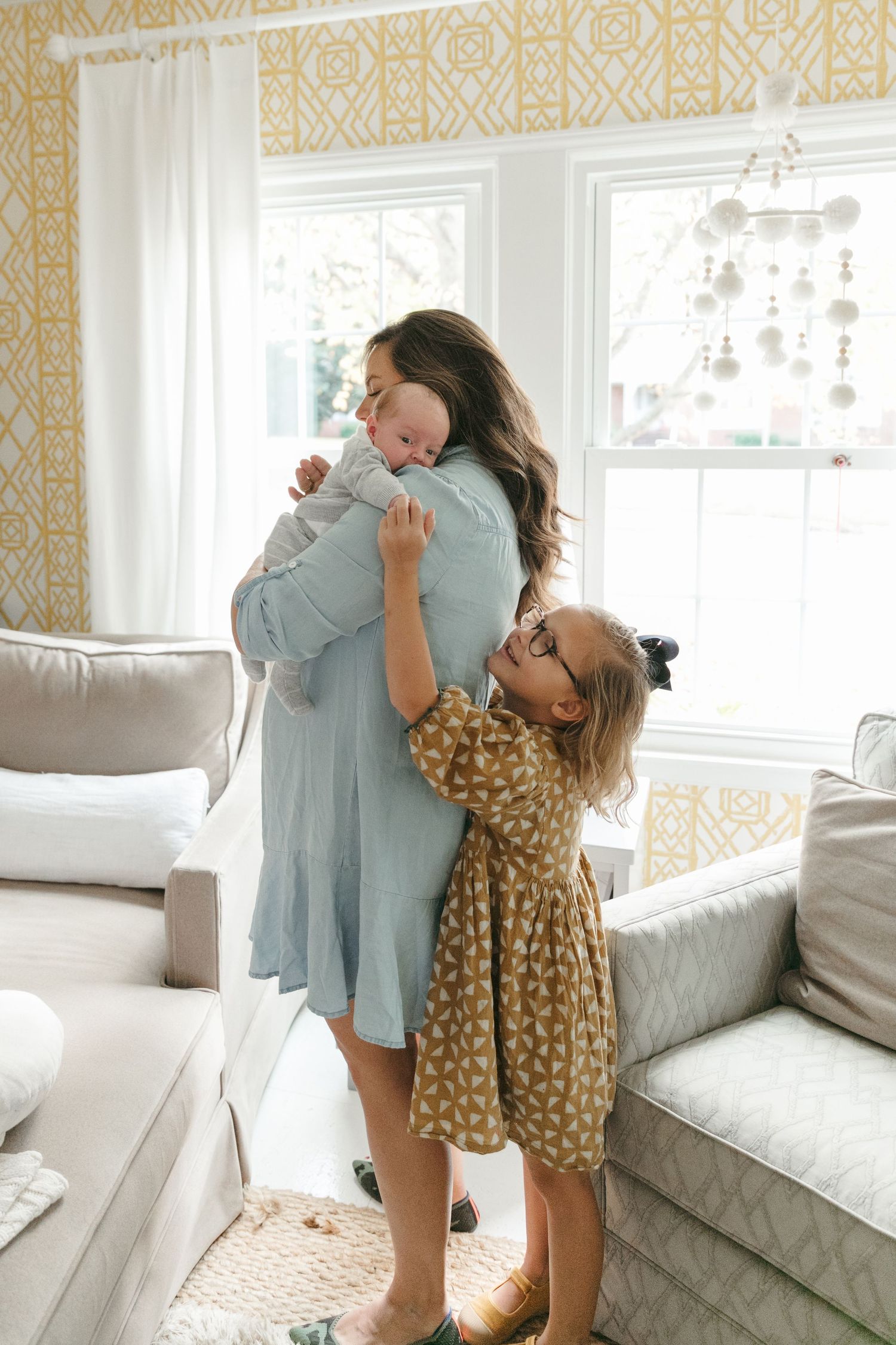Sweet family moment as child hugs parent holding newborn in sunny living room.