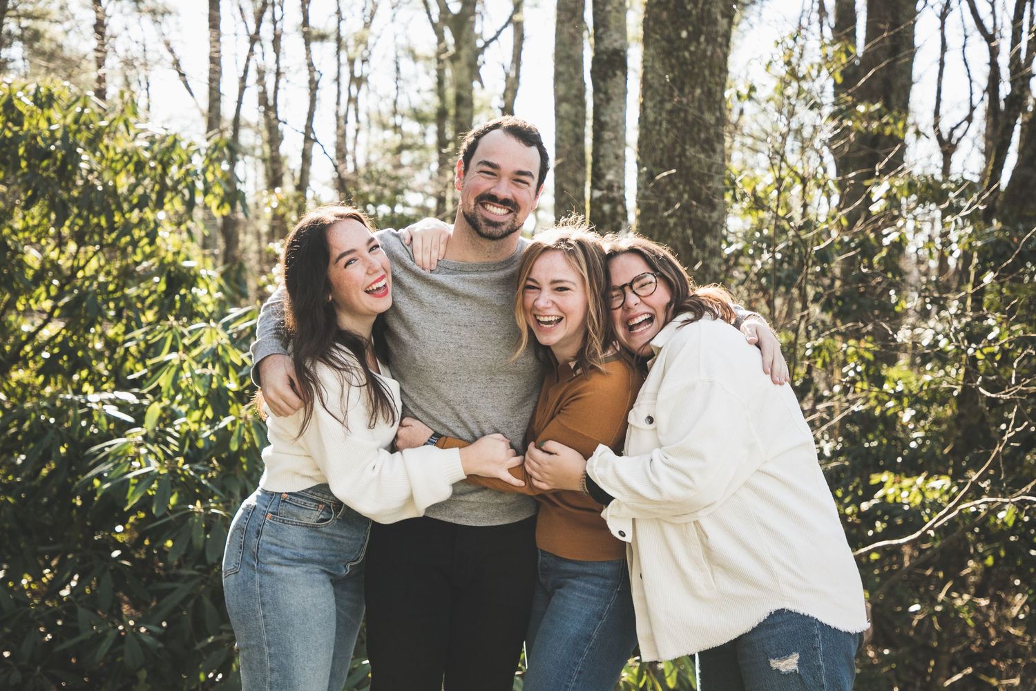 Group of people hugging and laughing together in a wooded outdoor setting.