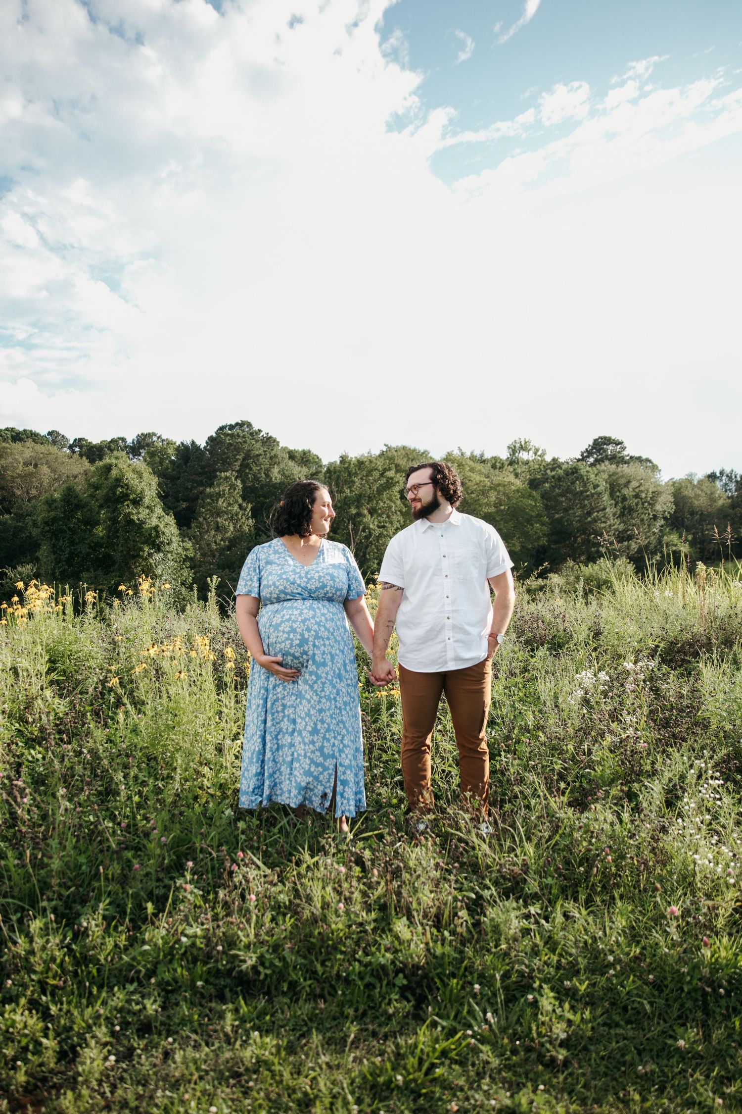 Couple holding hands in a summer meadow with blue sky and wildflowers.