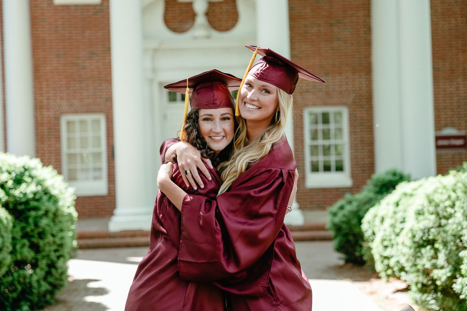 Two graduates in maroon caps and gowns embrace while celebrating on campus.