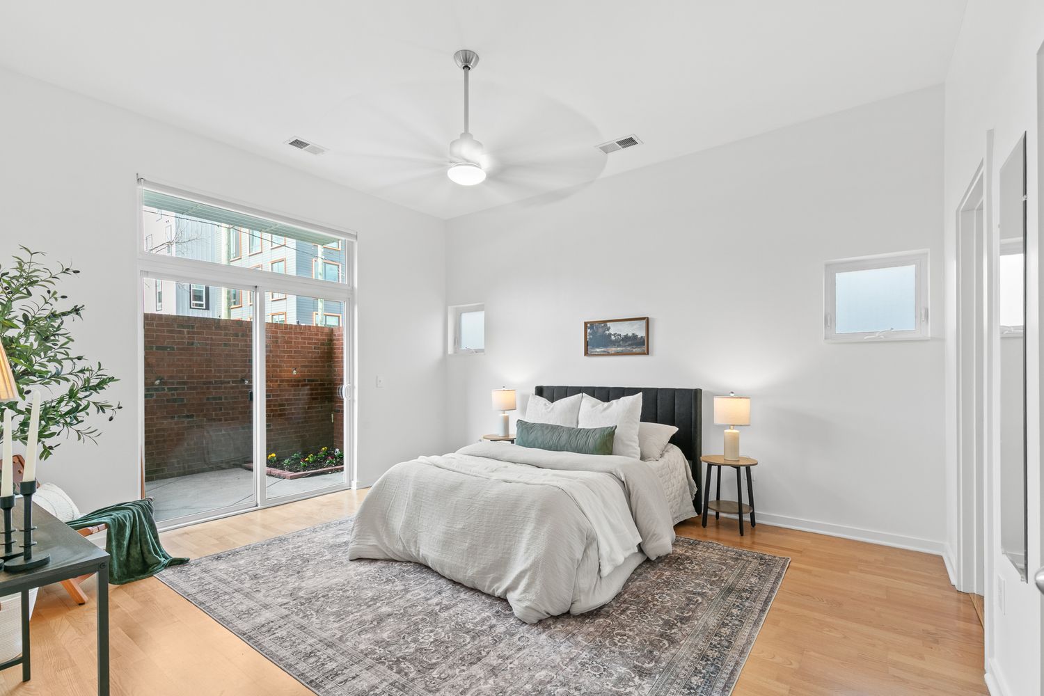 Bright modern bedroom with hardwood floors, ceiling fan, large patio doors, and a cozy bed with green accent pillows.