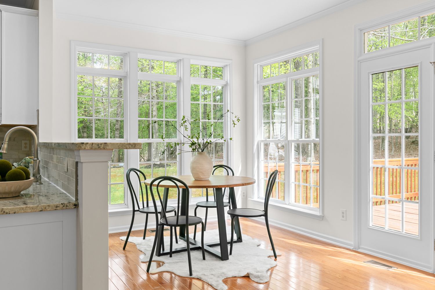 Bright sunlit dining nook with round wood table, black chairs, hardwood floors, and large windows overlooking lush greenery.