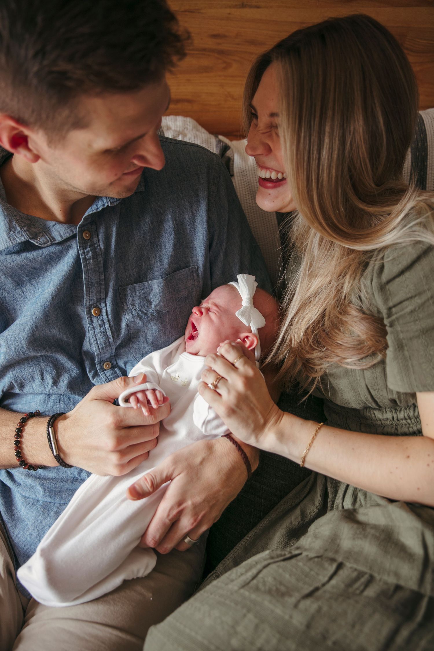 A couple shares a joyful moment with their laughing newborn baby on a couch.