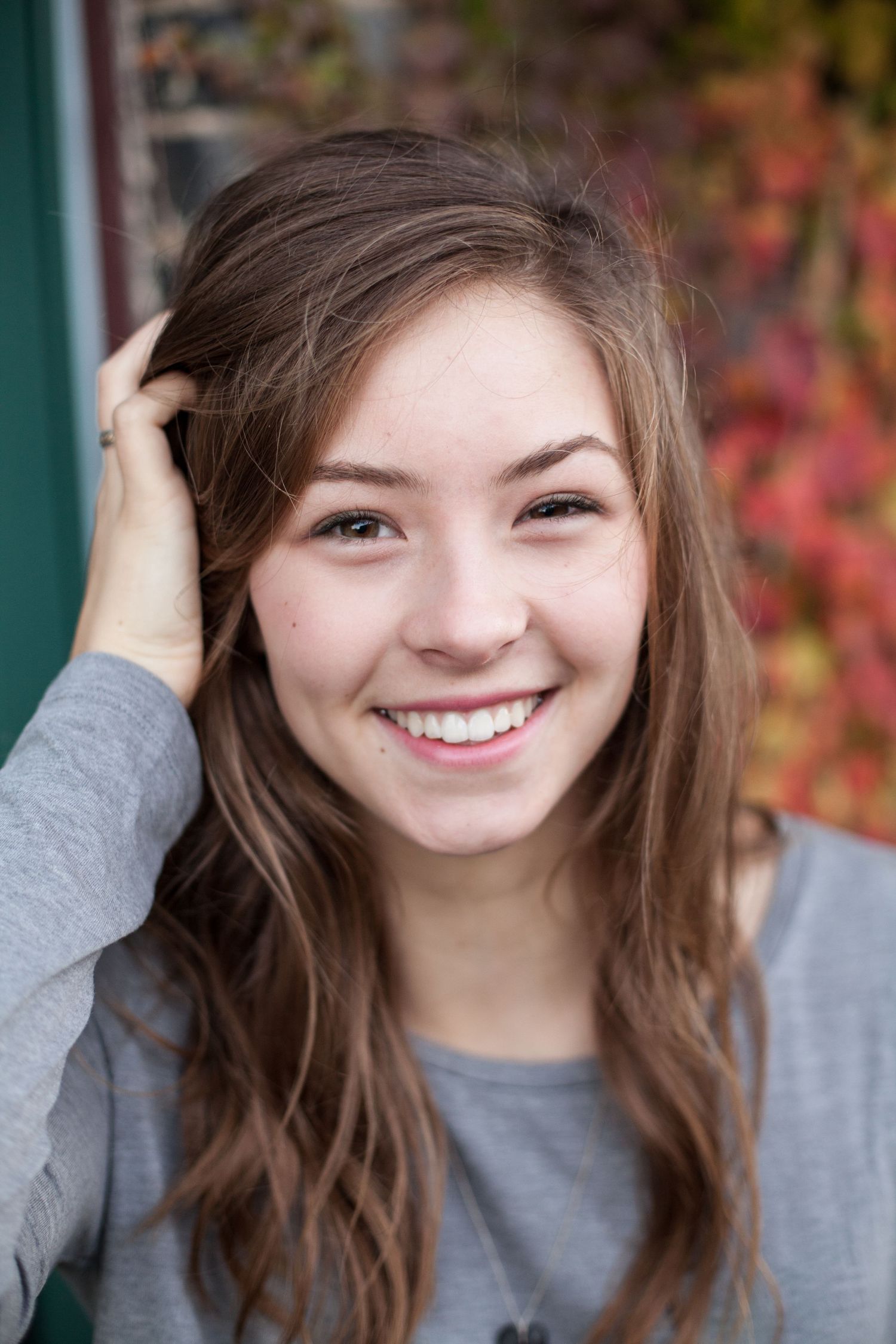 Portrait of a smiling person with long brown hair wearing a grey sweater against a background with autumn foliage.