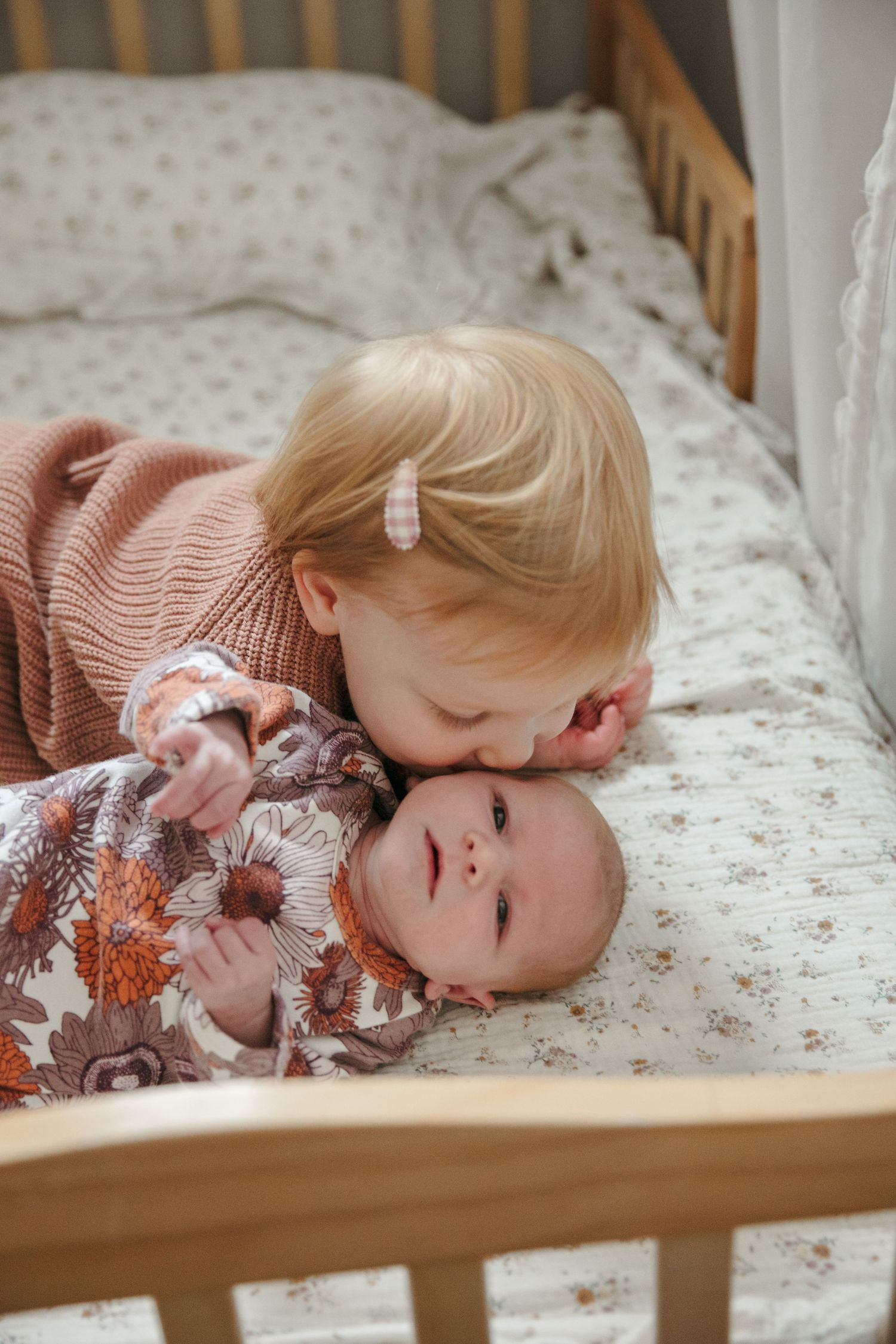 A toddler in pink sweater cuddles with baby sibling on patterned bedding in wooden crib.
