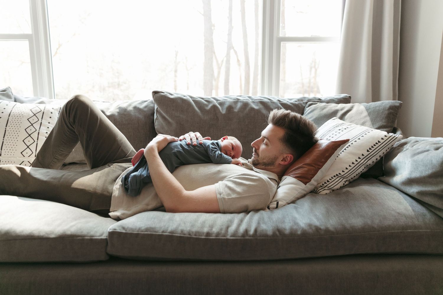 Relaxed moment on grey couch with sleeping infant resting on chest.
