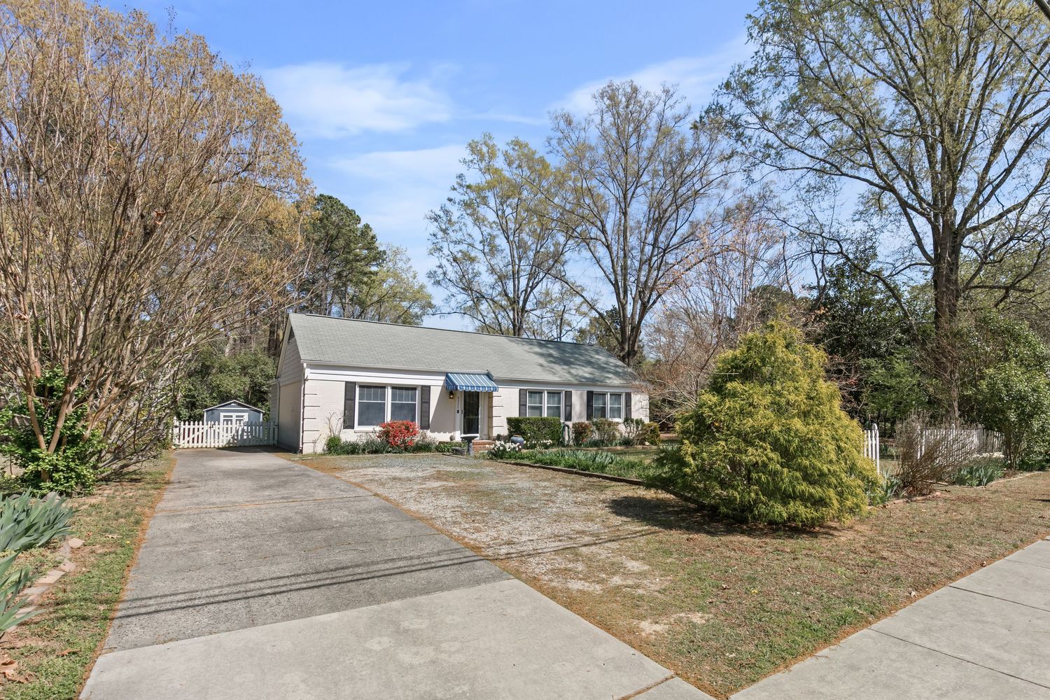Charming ranch-style home with gray roof, red front door, manicured shrubs, and a concrete driveway surrounded by mature trees.