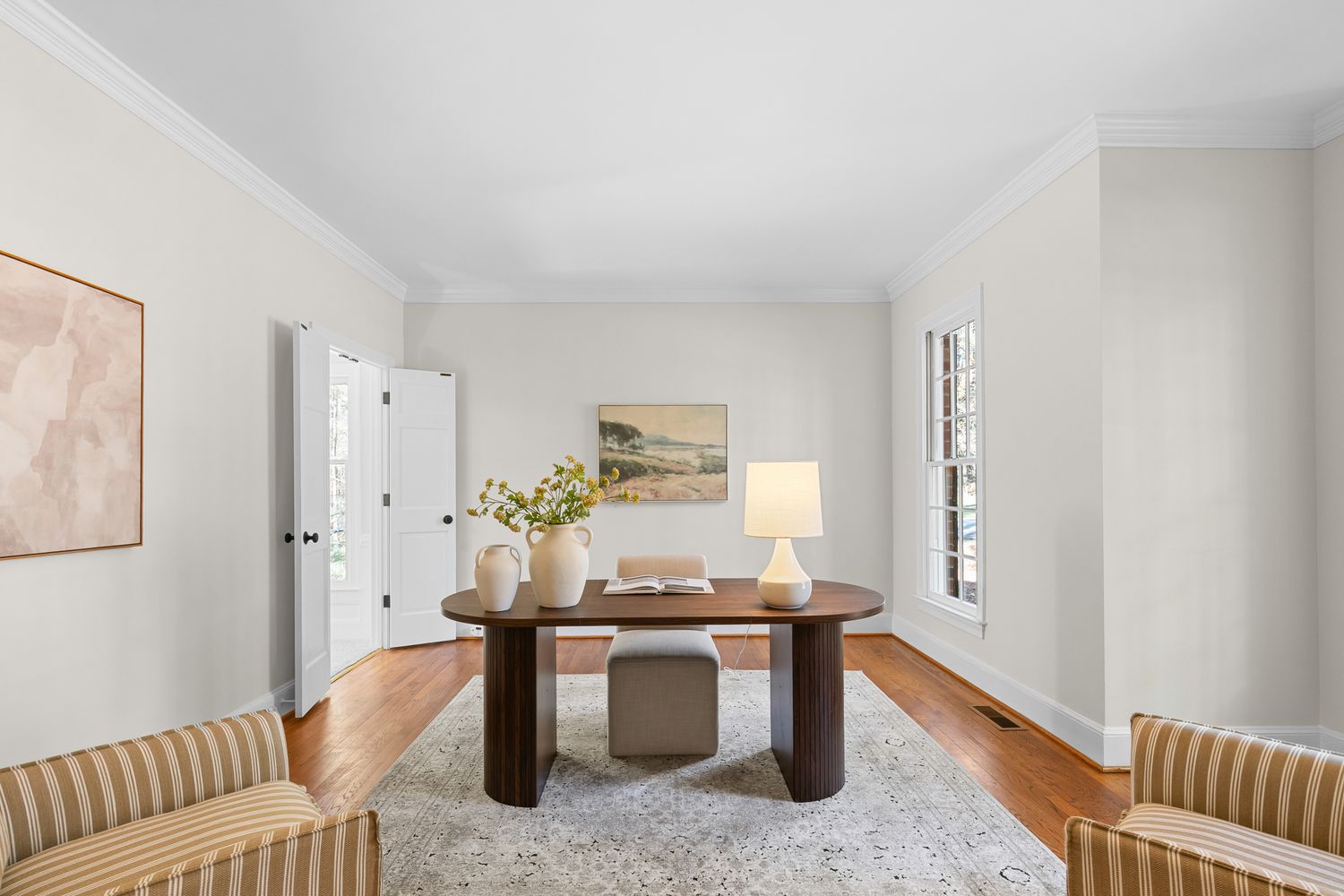 Elegant home office with round walnut desk, striped armchairs, hardwood floors, and neutral walls with crown molding.