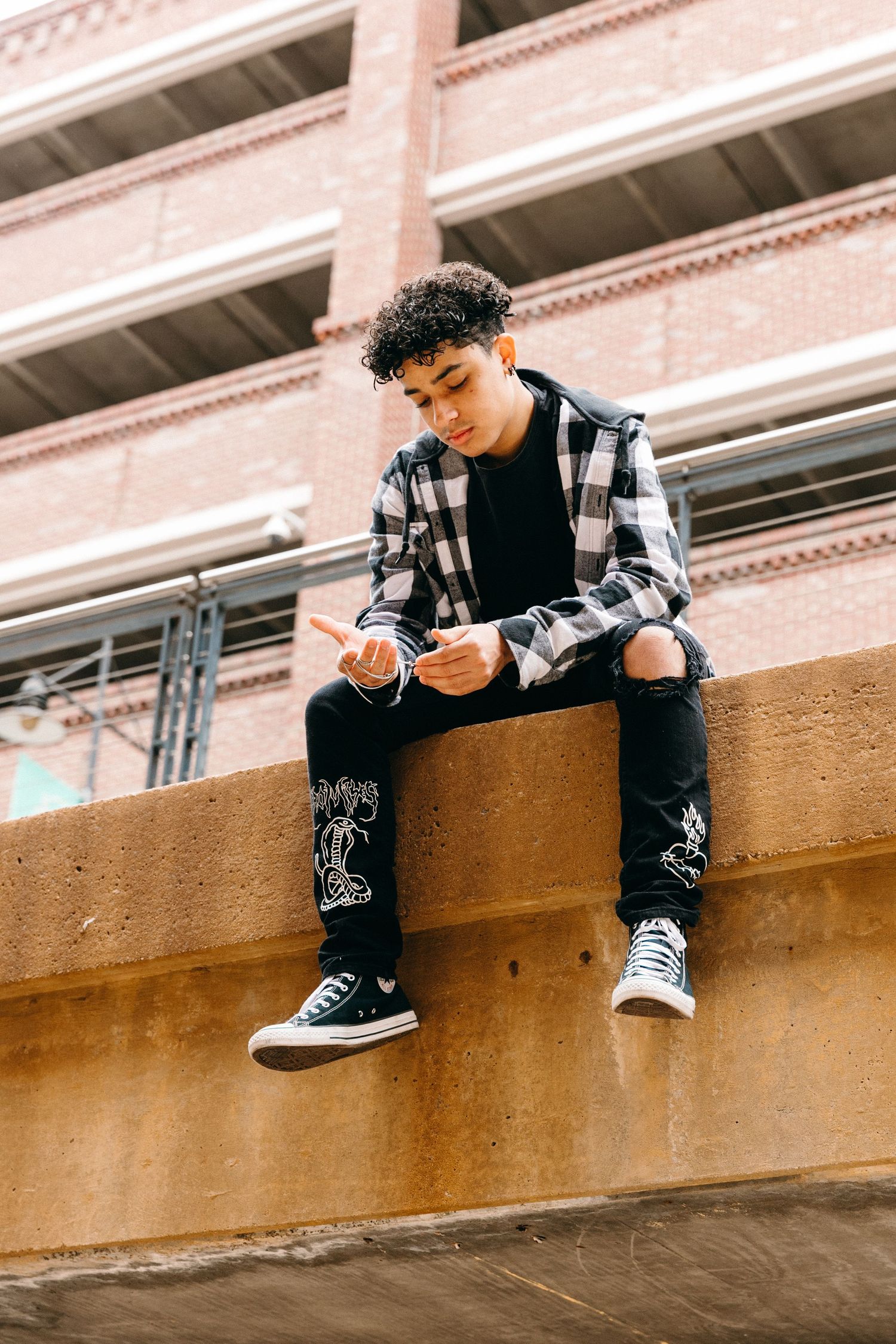 Person in plaid shirt and ripped jeans sitting on concrete ledge checking phone against urban backdrop.