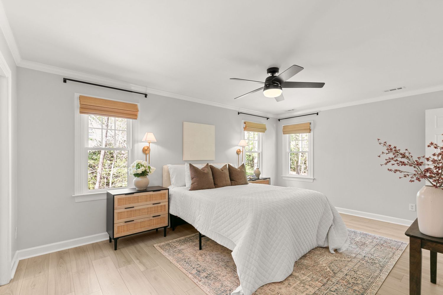 Bright bedroom with white bedding, ceiling fan, bamboo shades, hardwood floors, and a wooden dresser with floral decor.