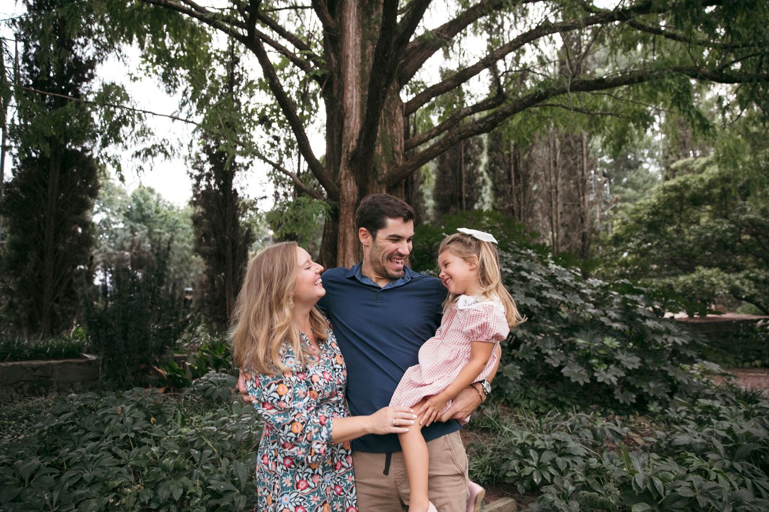 A loving family portrait taken in a lush garden setting with tall trees in the background.