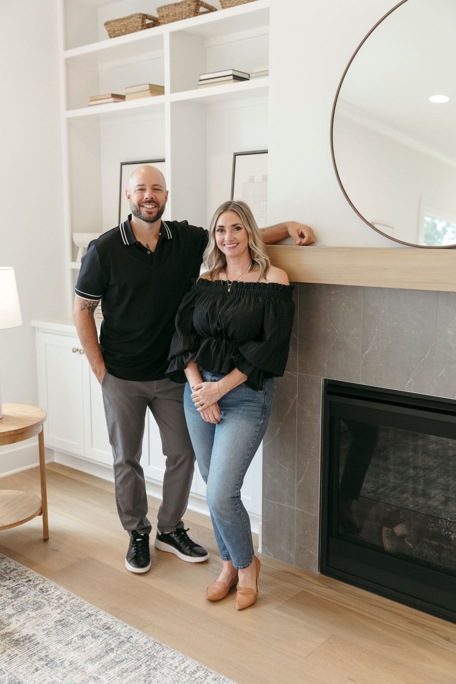 Casual couple standing by modern fireplace with built-in shelves and round mirror decor.