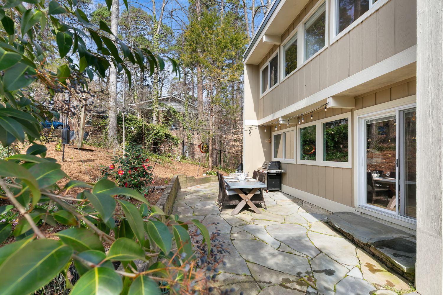 Stone patio with outdoor dining set beside a modern home surrounded by tall trees and lush greenery.