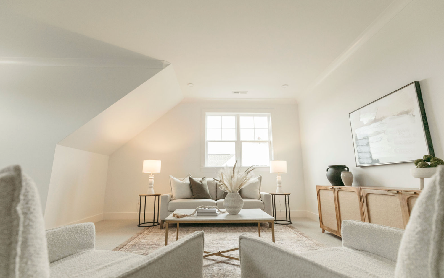 Bright attic living room with white armchairs, sofa, floor lamps, and wood accent furniture under sloped ceiling.