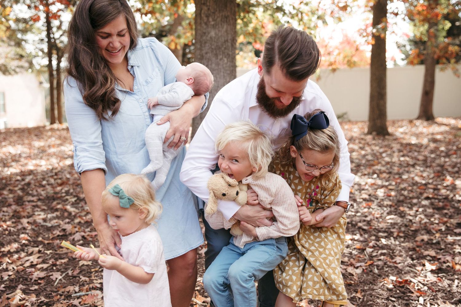 A loving family shares joyful laughter together during an autumn photoshoot in a wooded setting with fallen leaves.