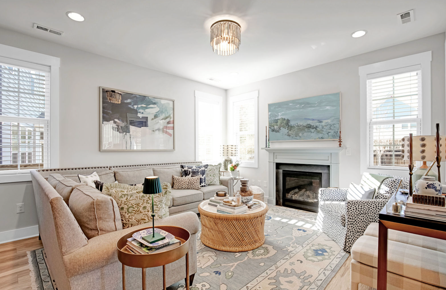 Bright living room with fireplace, neutral sofas, patterned rug, artwork, and natural light from large windows.