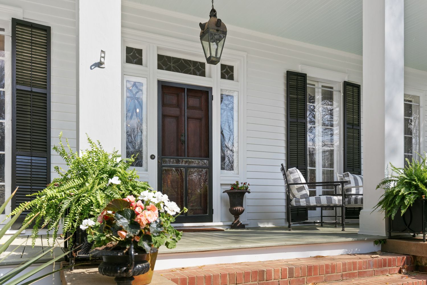 Elegant Southern home entrance with white columns, dark shutters, brick steps, and lush potted plants flanking a classic wooden door.