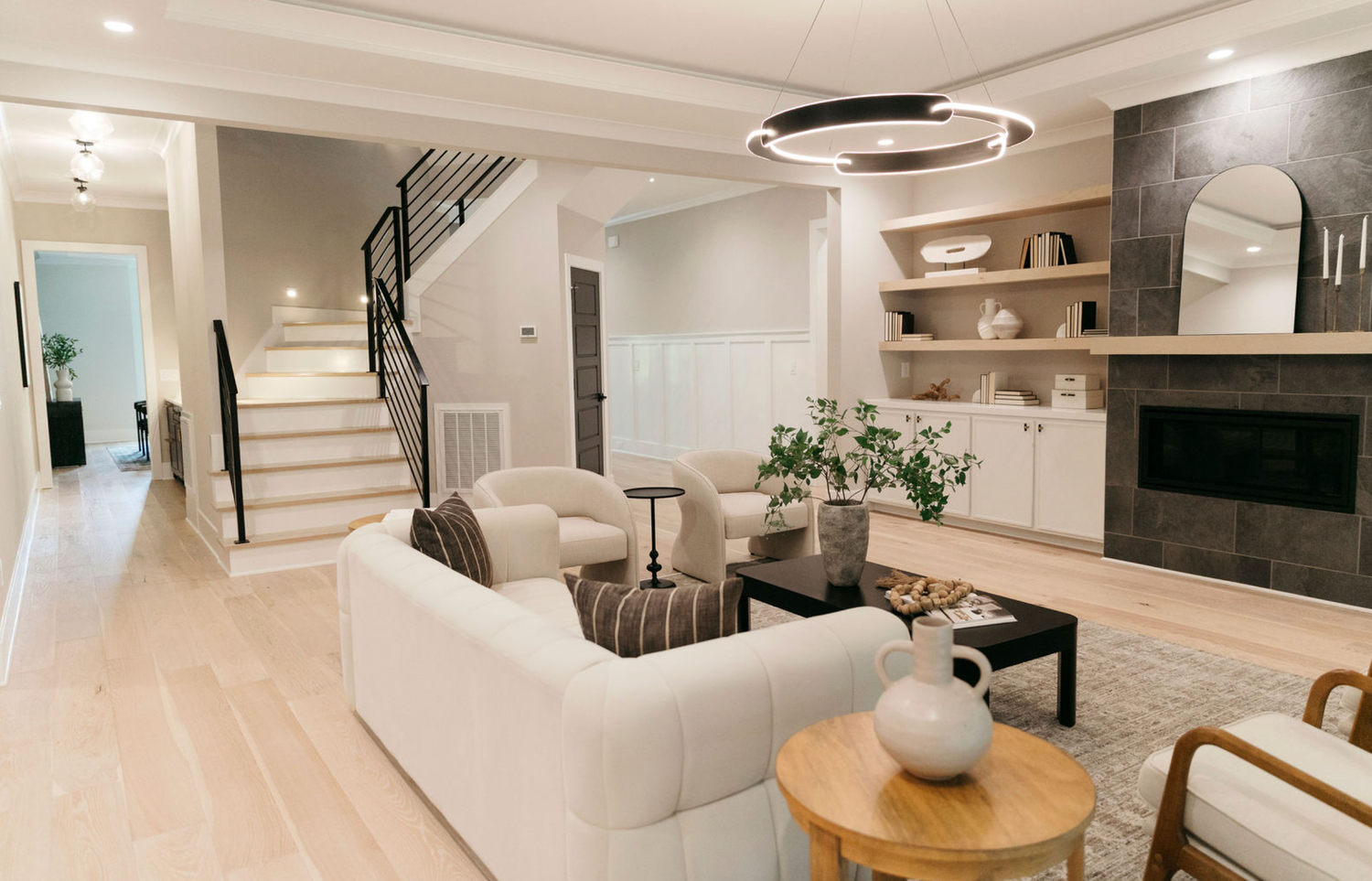 Modern open-concept living room with white sofa, built-in shelves, stone fireplace, circular chandelier, and light hardwood floors.