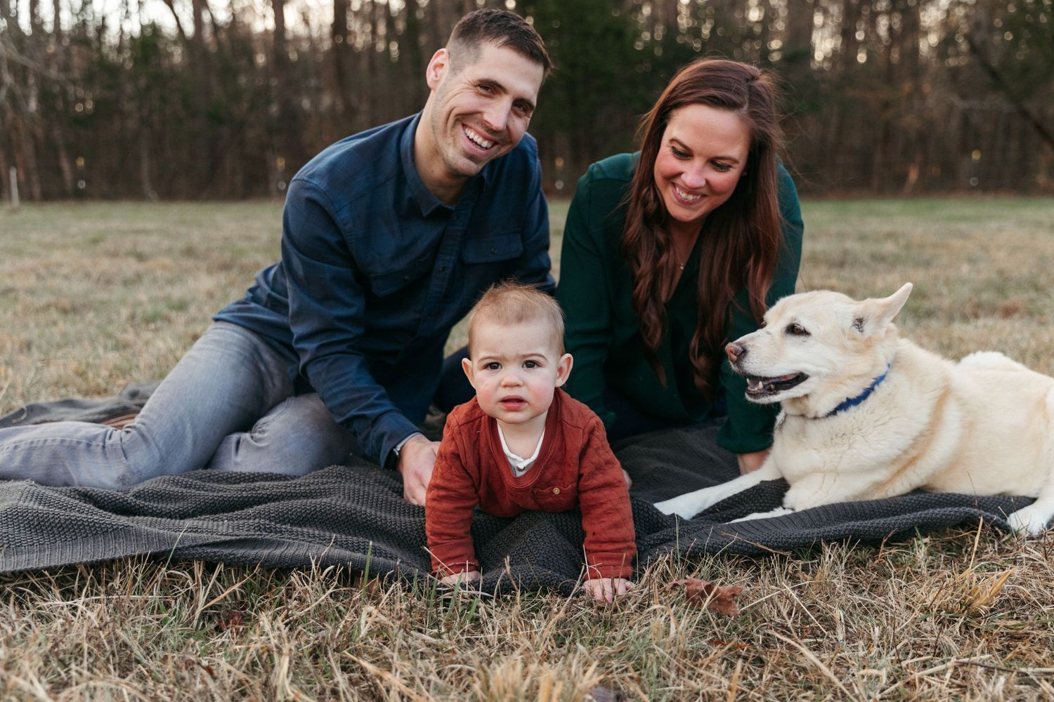 Family and their white dog share a loving moment while relaxing on a blanket in a grassy field.