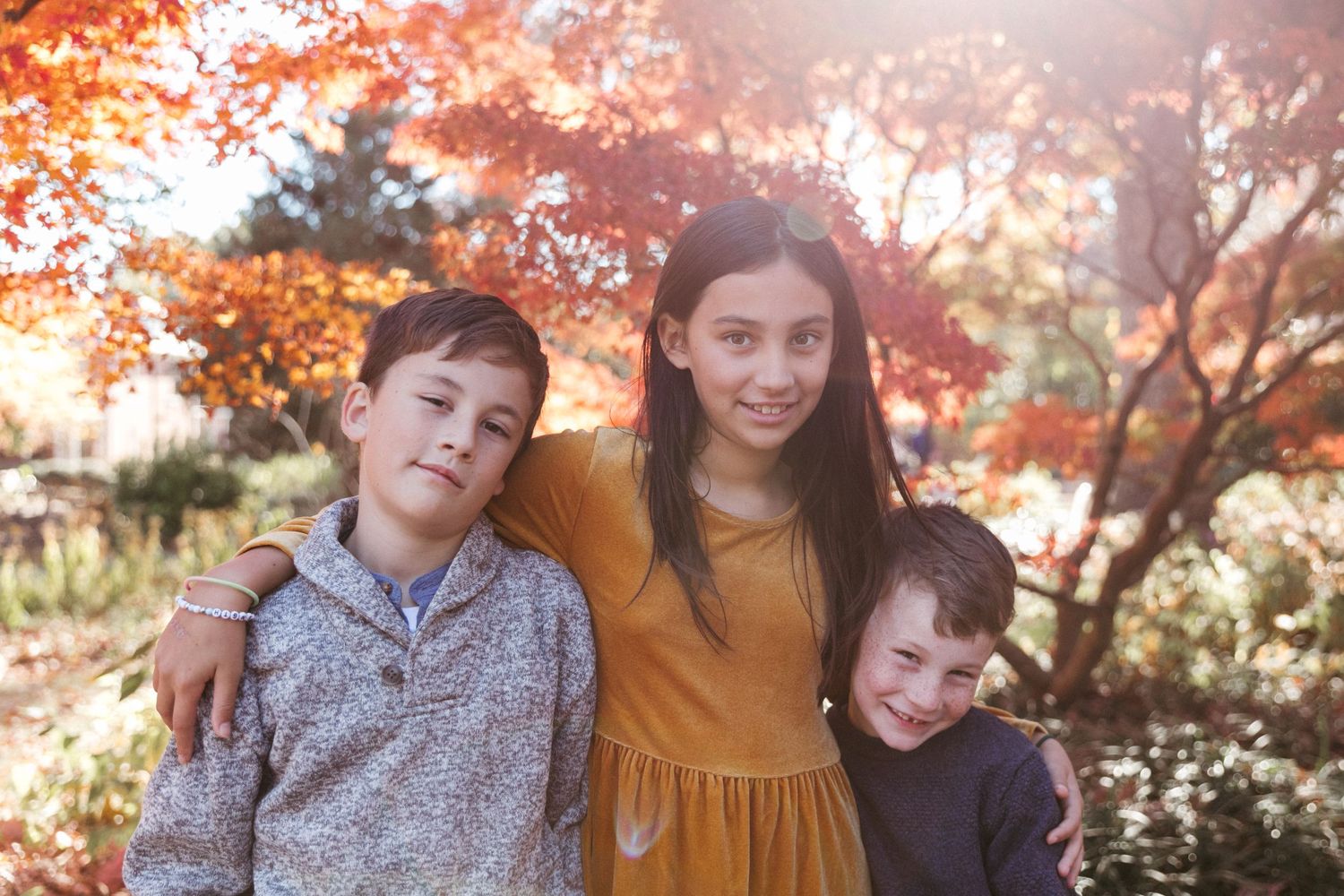 Three young people pose together under vibrant autumn foliage with warm orange tones in the background.