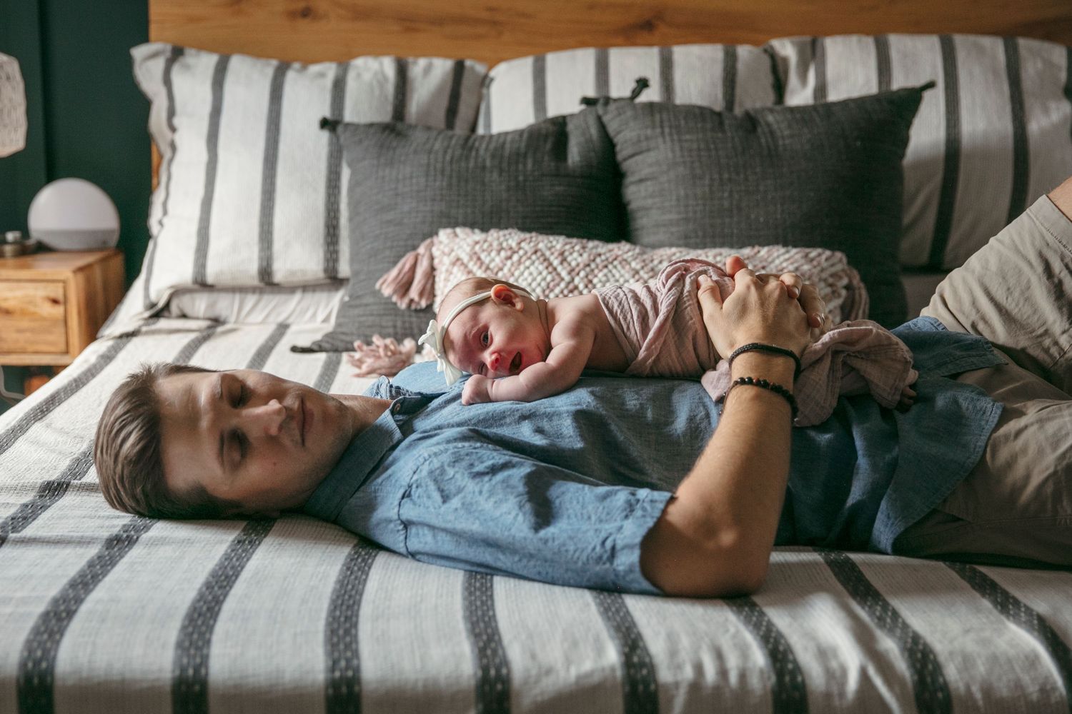 Bedroom interior with wooden bed frame against dark green wall and natural light. Father lays on bed with newborn baby.
