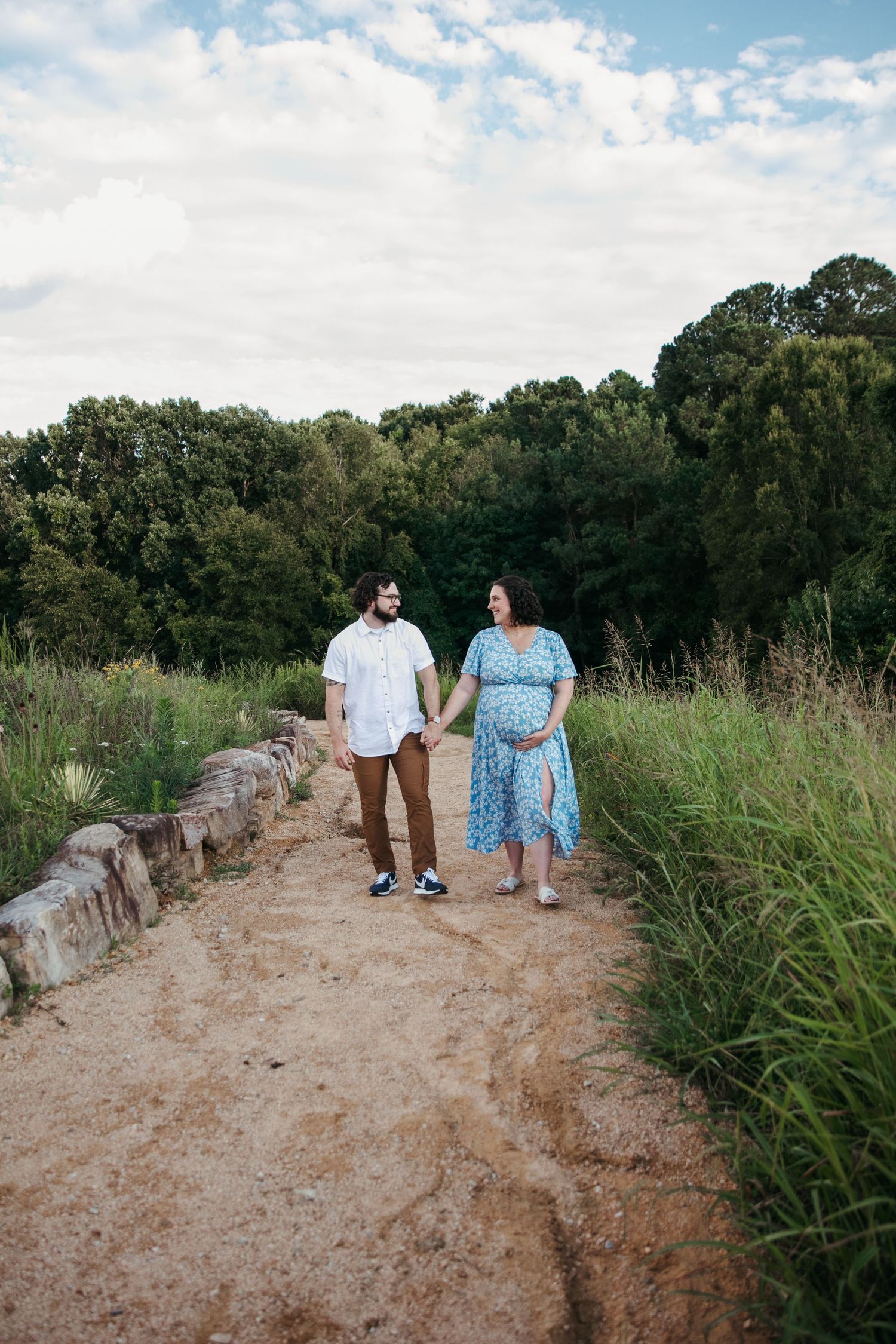 A couple walking along a rustic stone path surrounded by tall grass and trees on a sunny day.