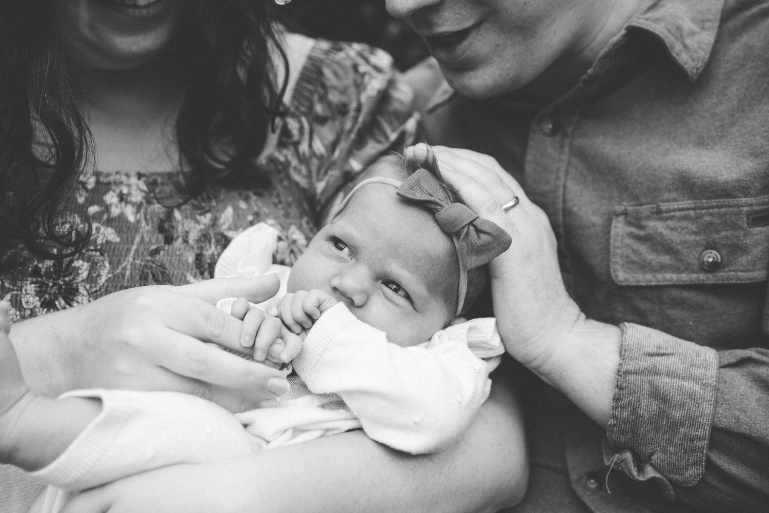 Black and white photo showing a small baby in white clothing being held and comforted by parents.