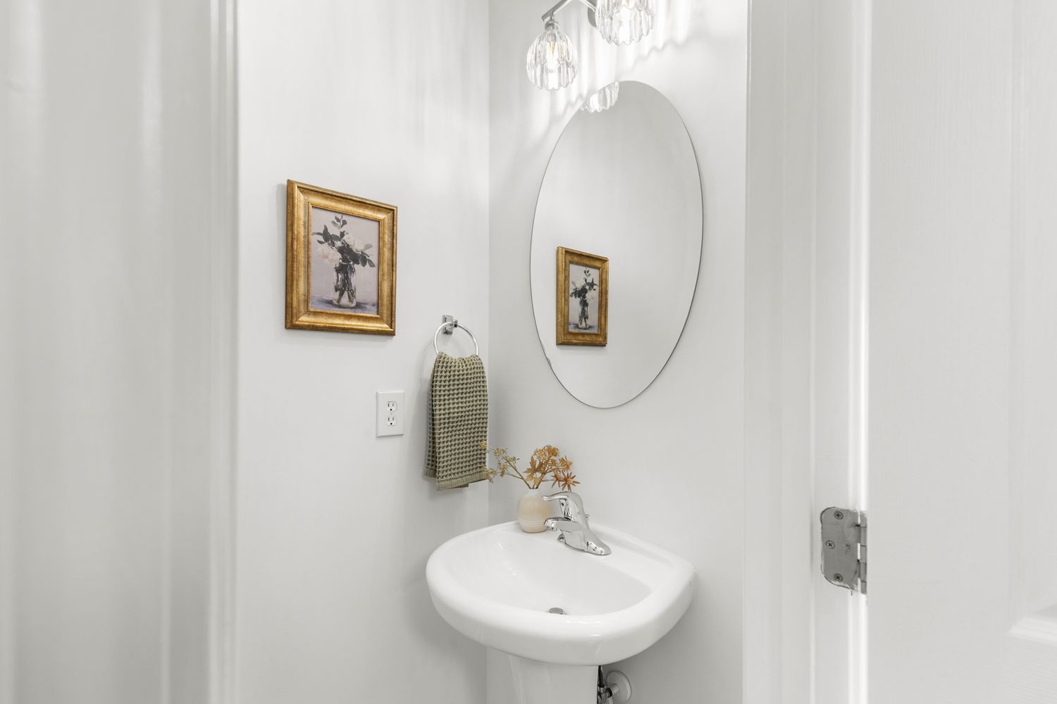 Elegant white powder room with pedestal sink, oval mirror, crystal chandelier, and gold-framed artwork.