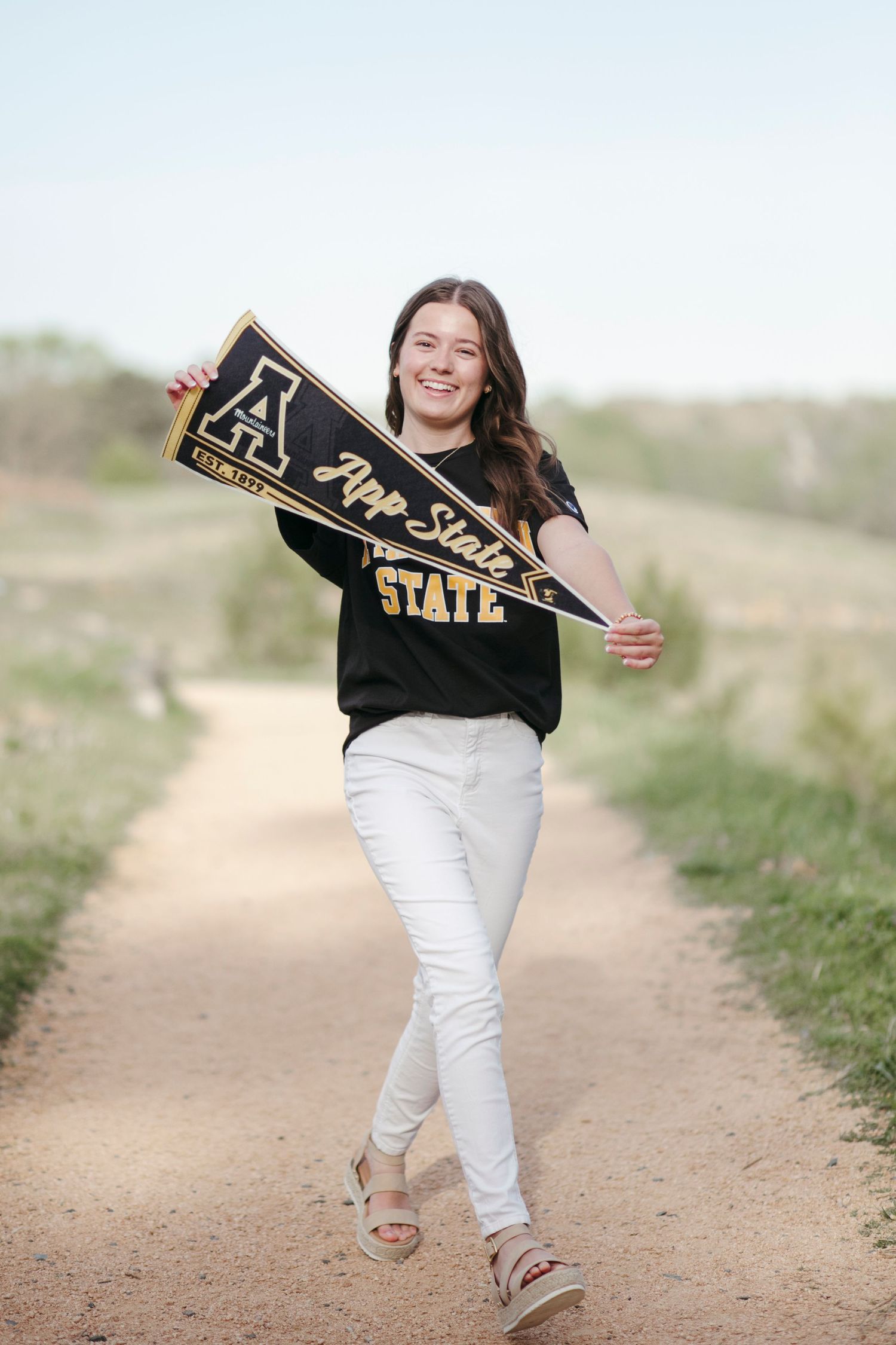 Student wearing black shirt and white pants holds App State pennant on dirt path.