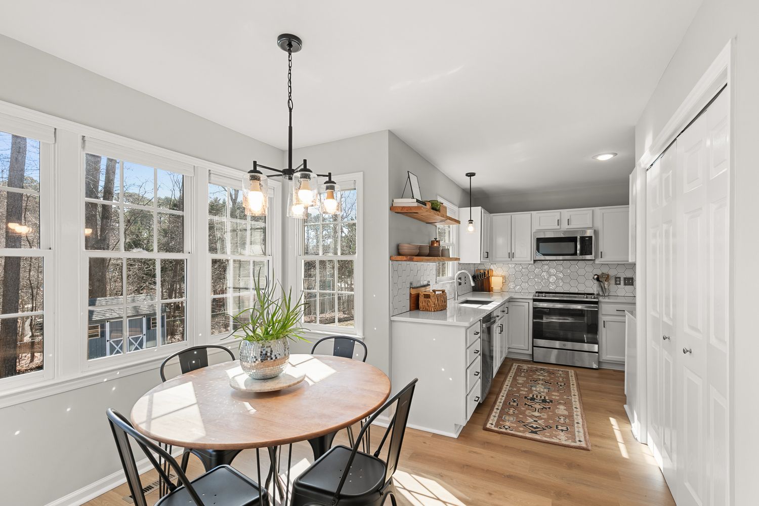 Bright dining area with round wood table, black chairs, large windows, open shelving, and modern kitchen with stainless appliances.