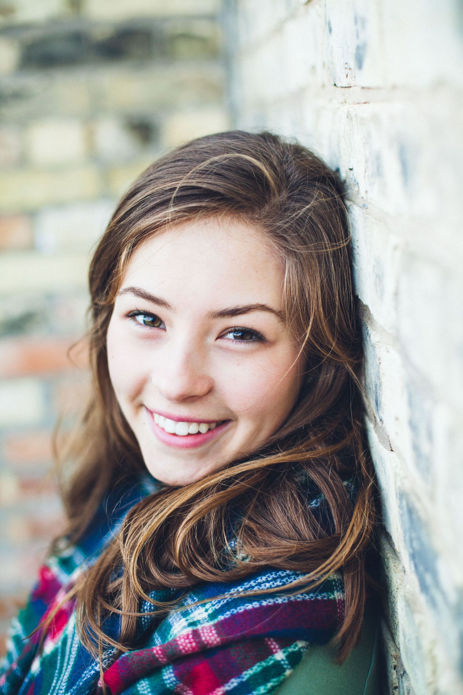 A person wearing a plaid scarf leans against a brick wall with a warm smile.