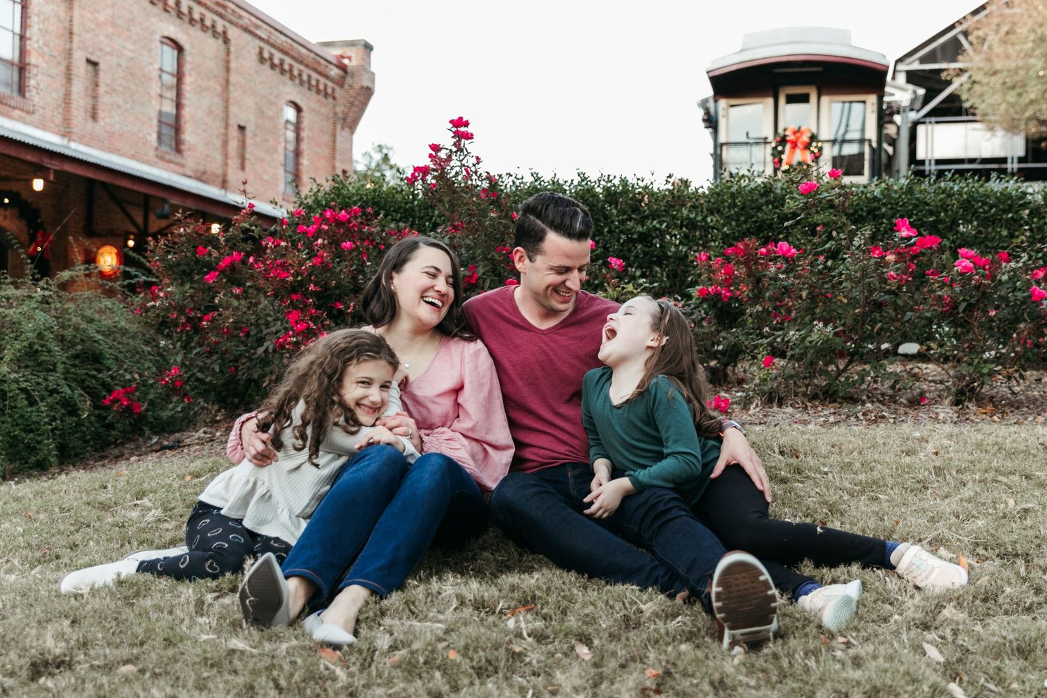 Family relaxing together on grass with red roses and brick building in background.