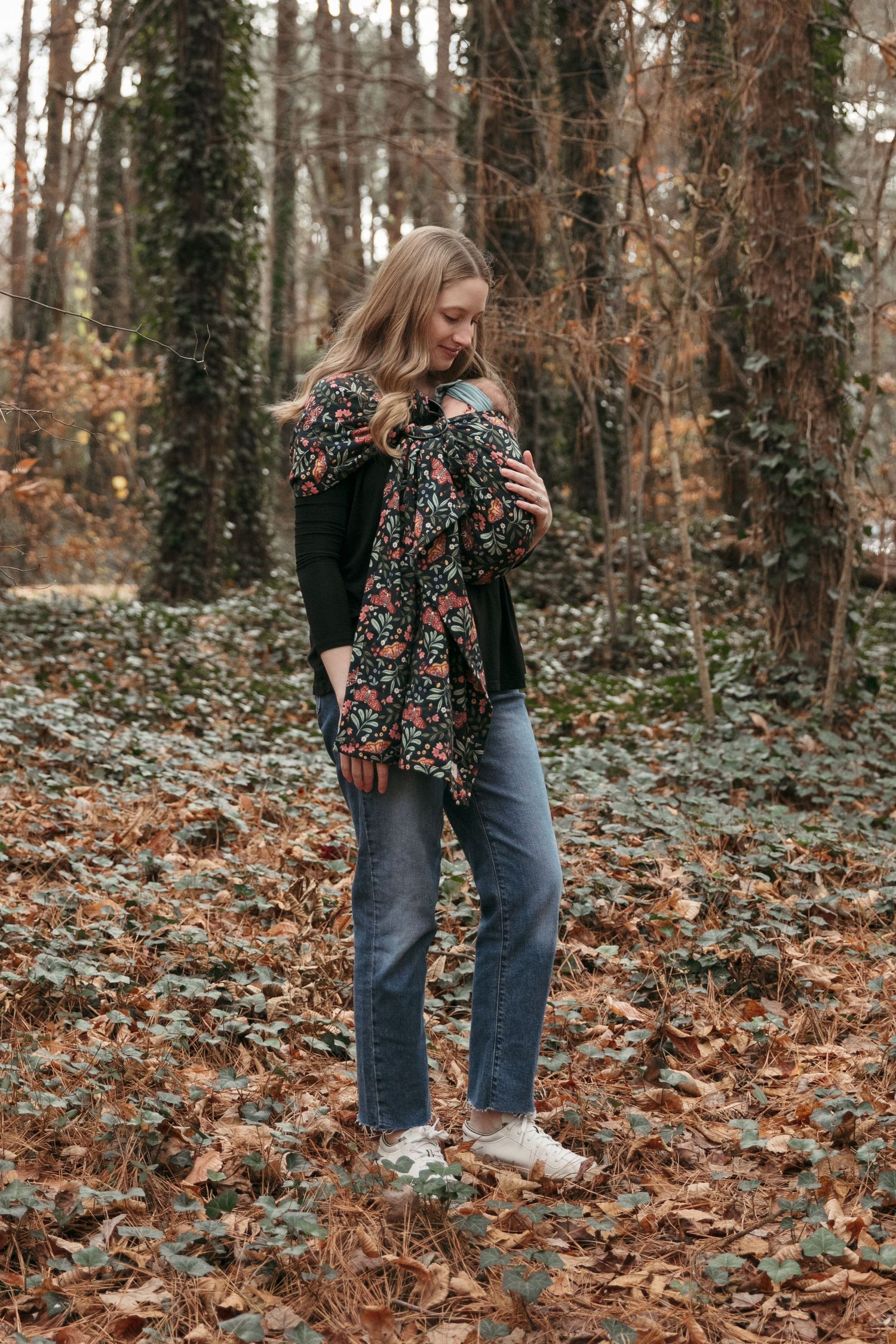 Person in floral top and jeans walks through autumn forest with fallen leaves covering the woodland floor.