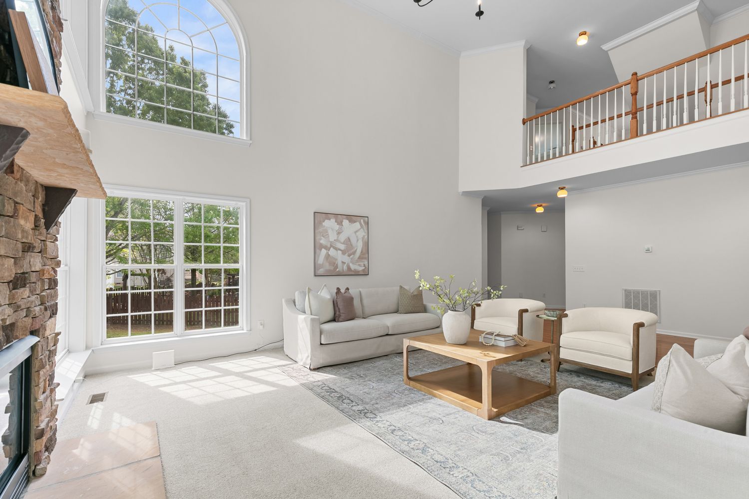 Bright two-story living room with white sofas, wood coffee table, stone fireplace, large windows, and open balcony above.