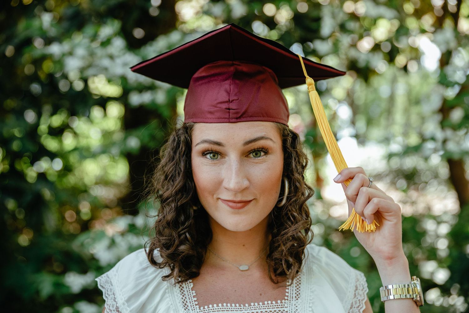 A graduation photo featuring someone in a maroon cap and gown with gold tassel against a blurred natural background.