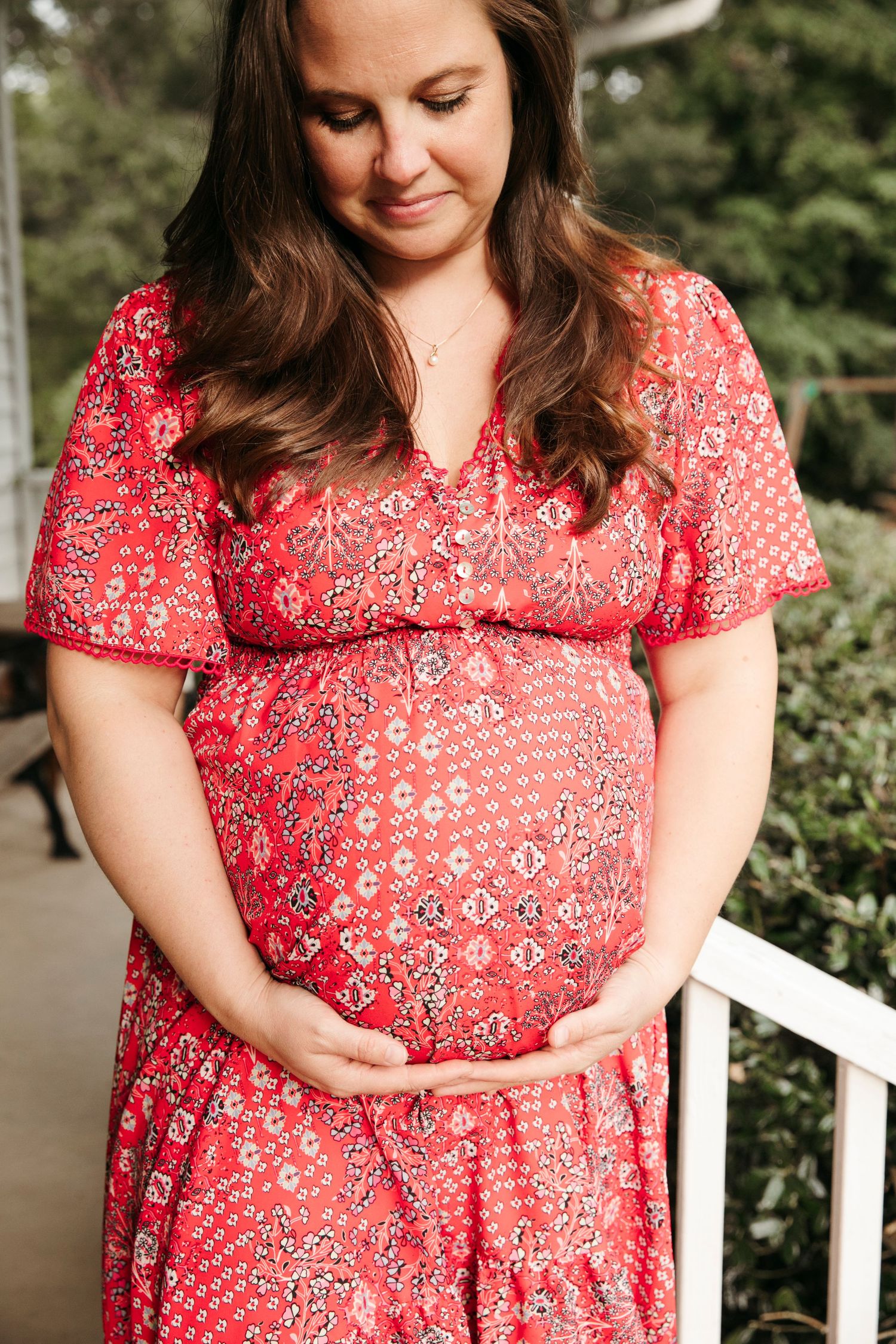 Close-up of woman in red floral dress on white porch during maternity photoshoot.
