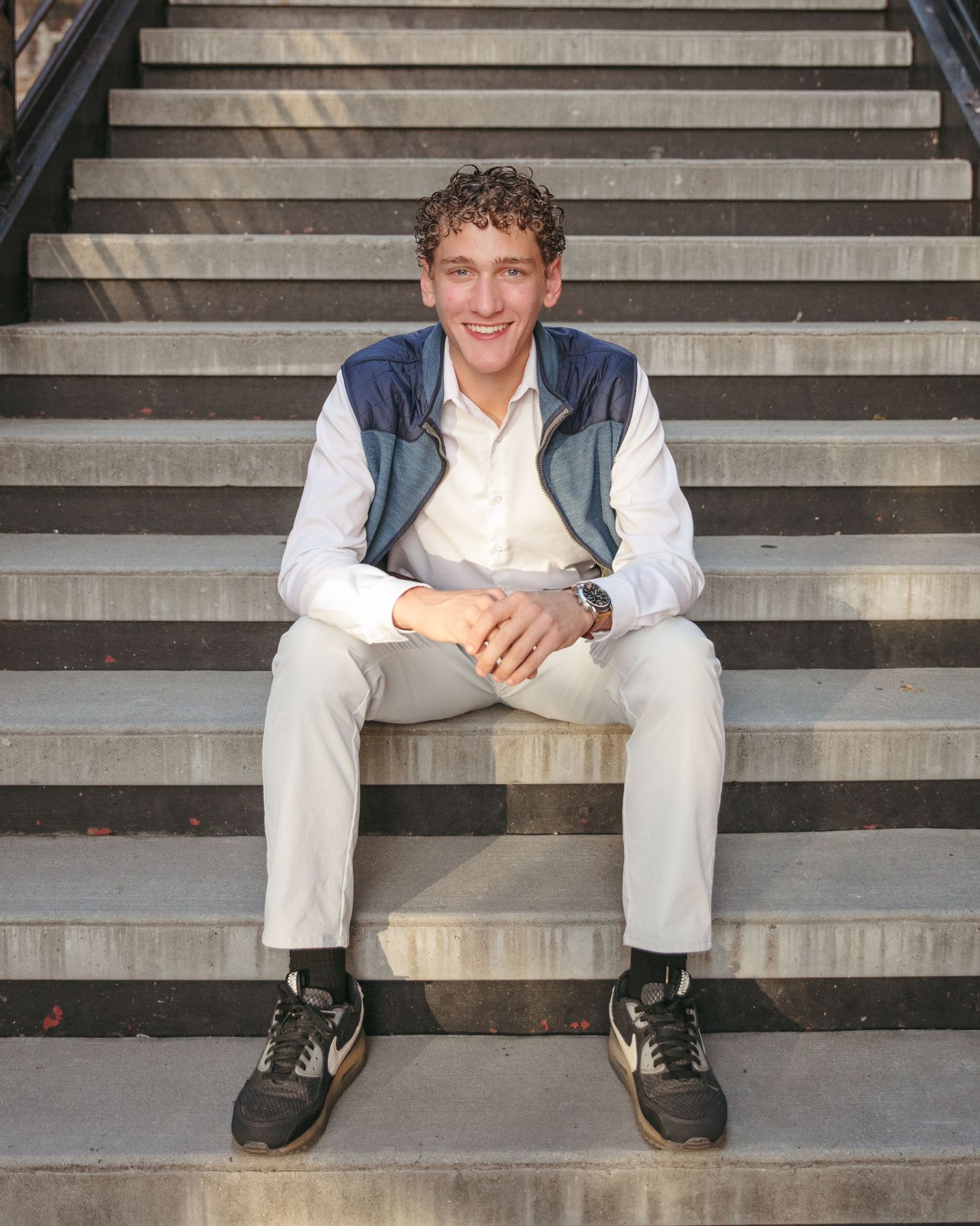 A casual portrait of someone seated on concrete stairs wearing a navy vest and white shirt.