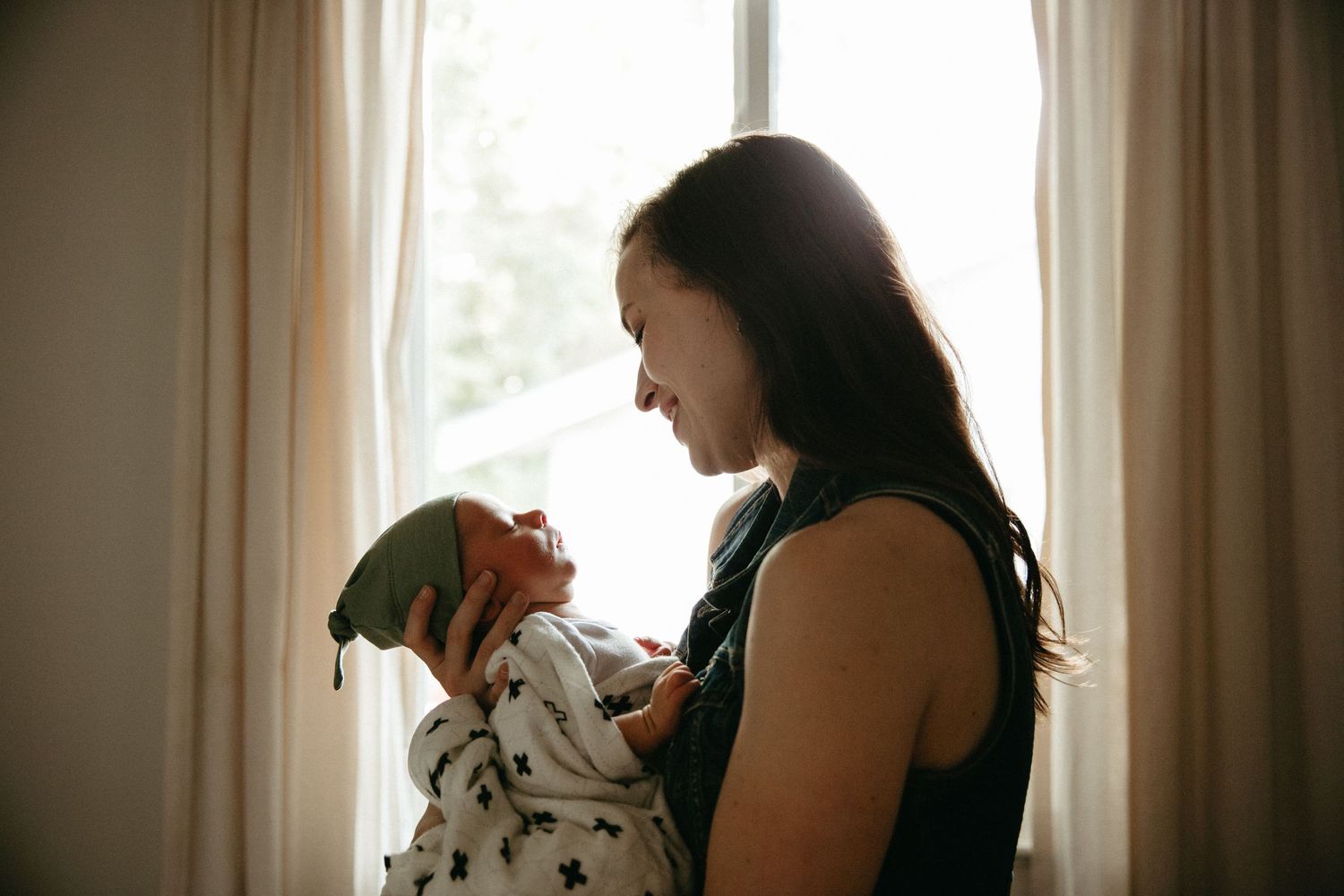 A parent cradles and lovingly looks down at their newborn baby by a window.