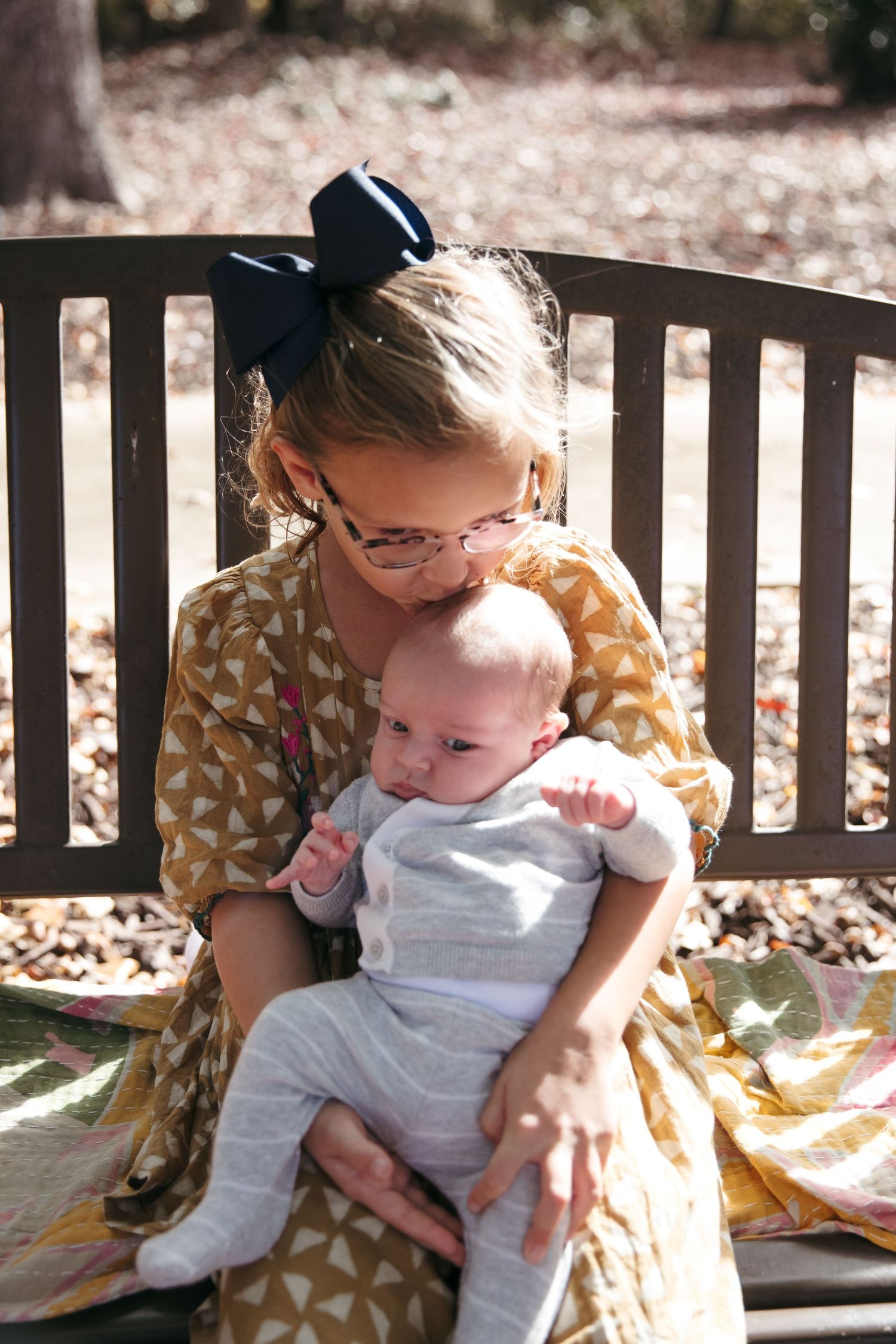 A baby in white overalls sits on a wooden bench with someone in a patterned dress on an autumn day.