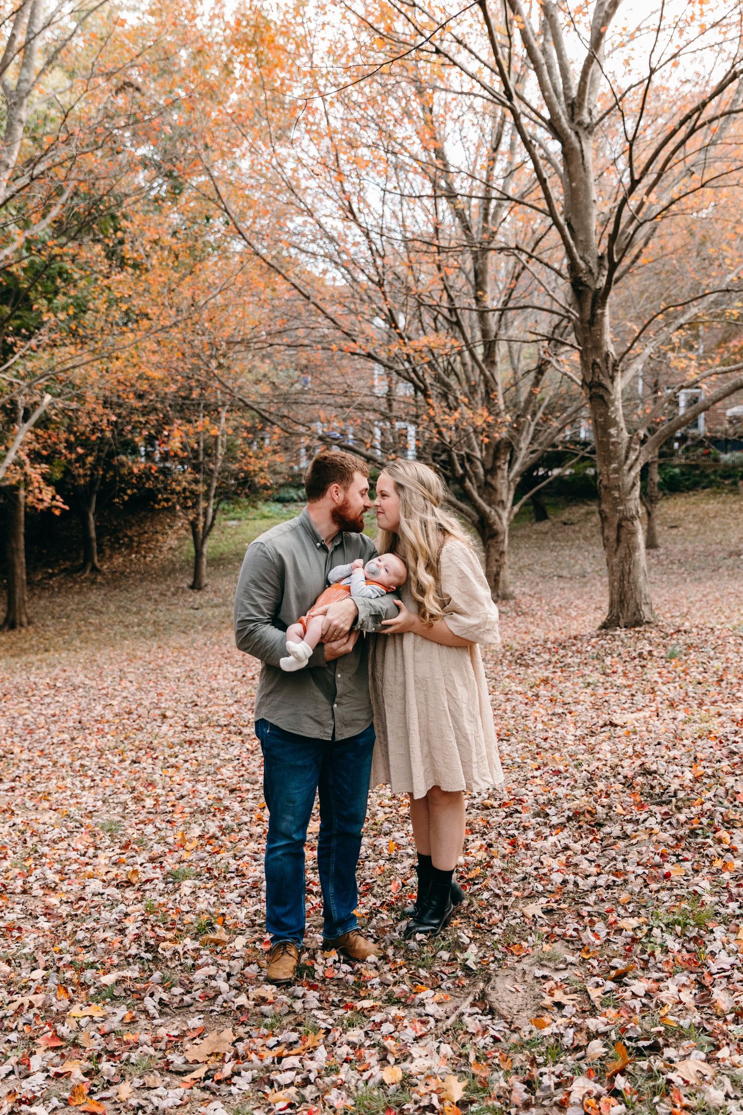 Couple sharing tender moments in autumn park with fallen leaves as baby sleeps in carrier.