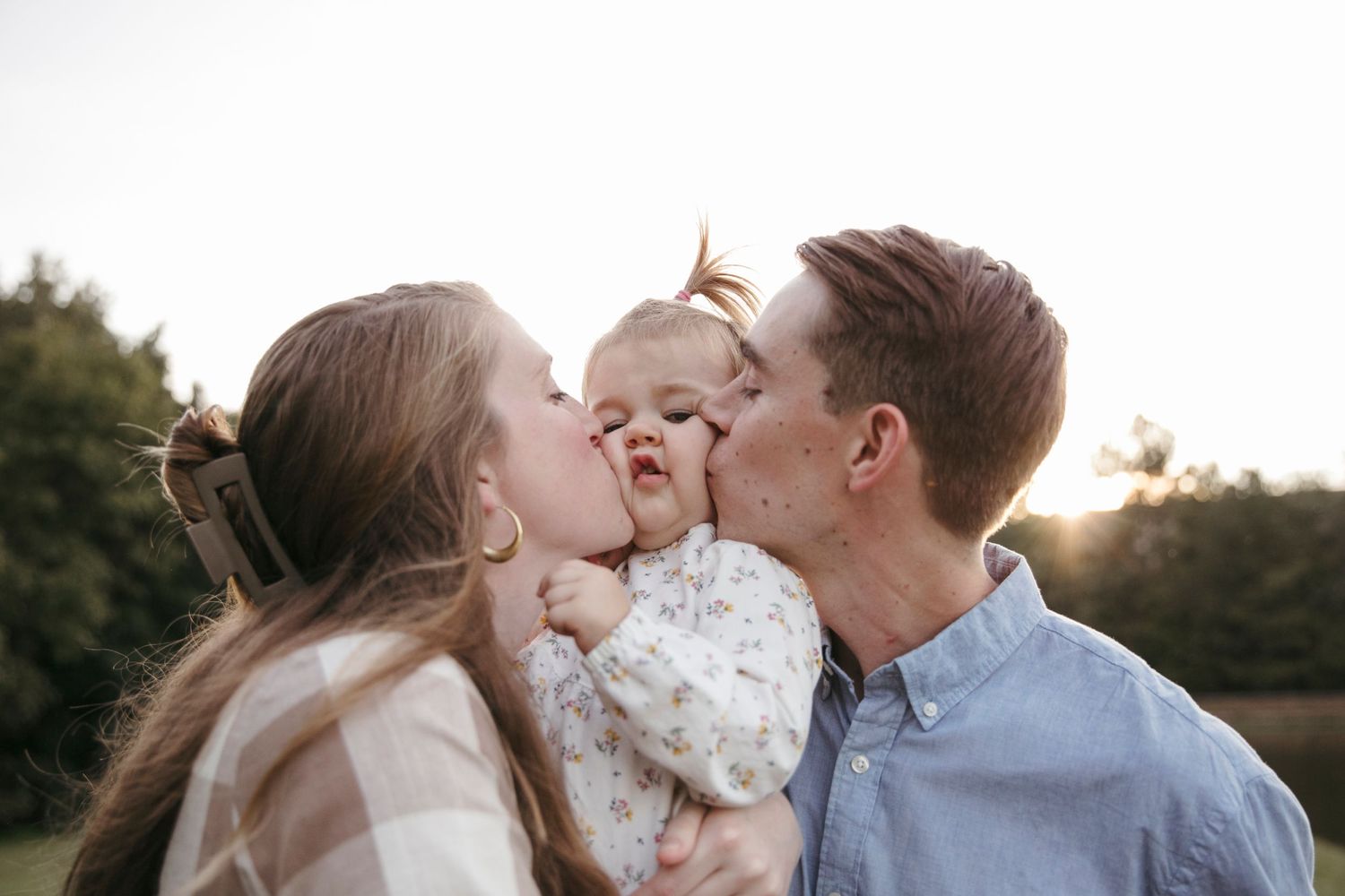 Parents sharing sweet kisses with their baby in a tender outdoor moment.
