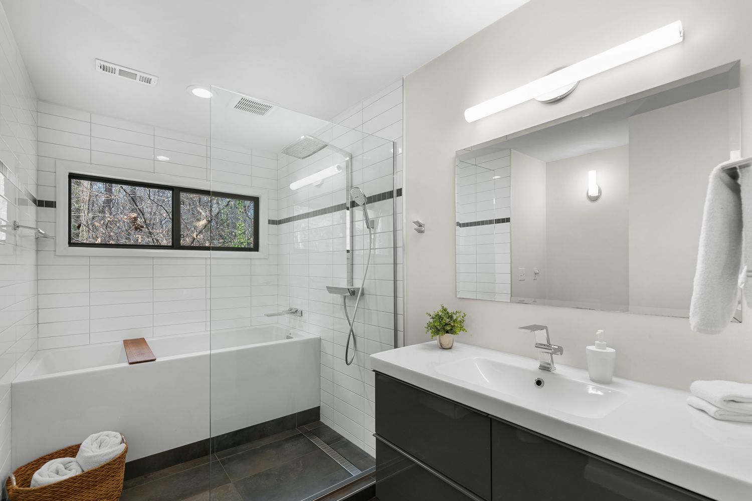 Modern bathroom with white subway tile, glass shower, soaking tub, dark vanity, double sinks, and large mirror.
