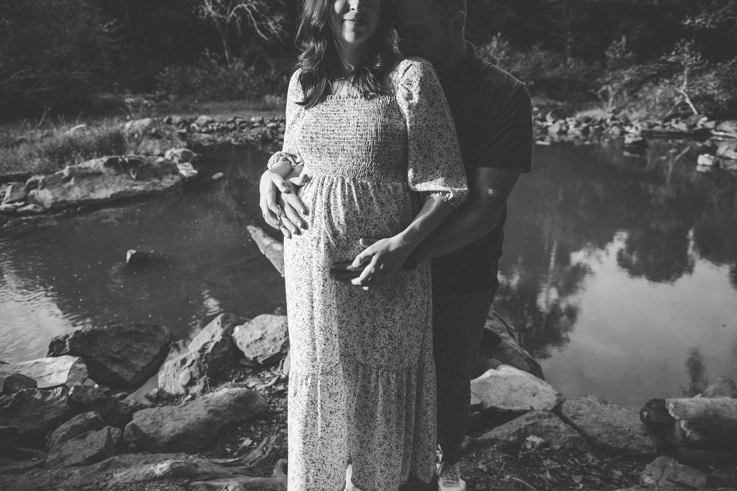 Black and white maternity photo with hands tenderly holding a pregnant belly by a lake.
