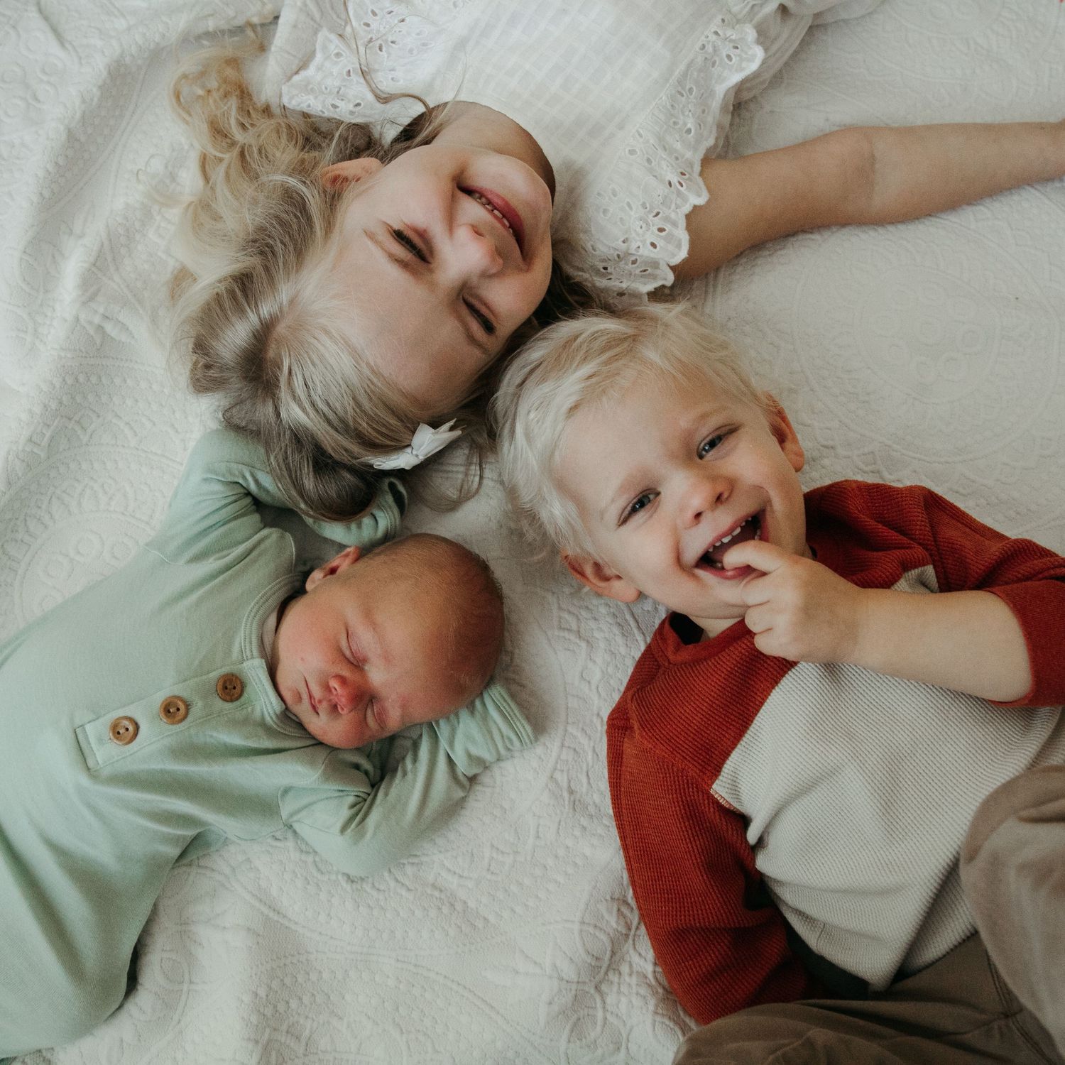Joyful family scene with children lying together on white bedding showing natural expressions.