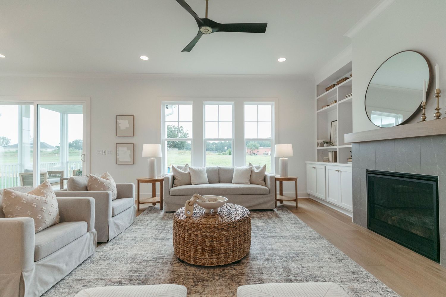 Modern coastal living room with white decor, rattan ottoman, built-in shelving and a black fireplace.