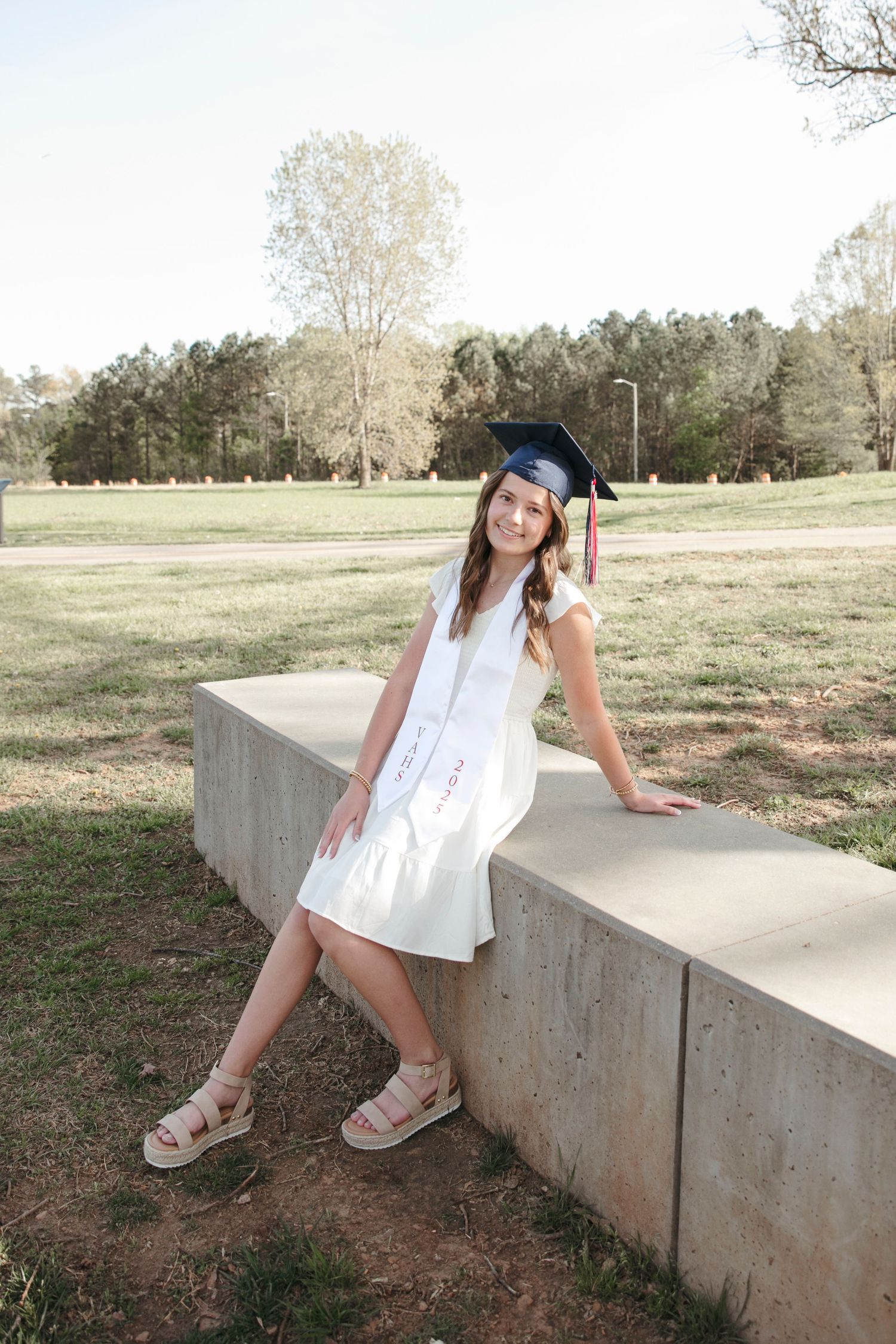 Graduate in white dress and blue cap poses on concrete wall in outdoor setting.