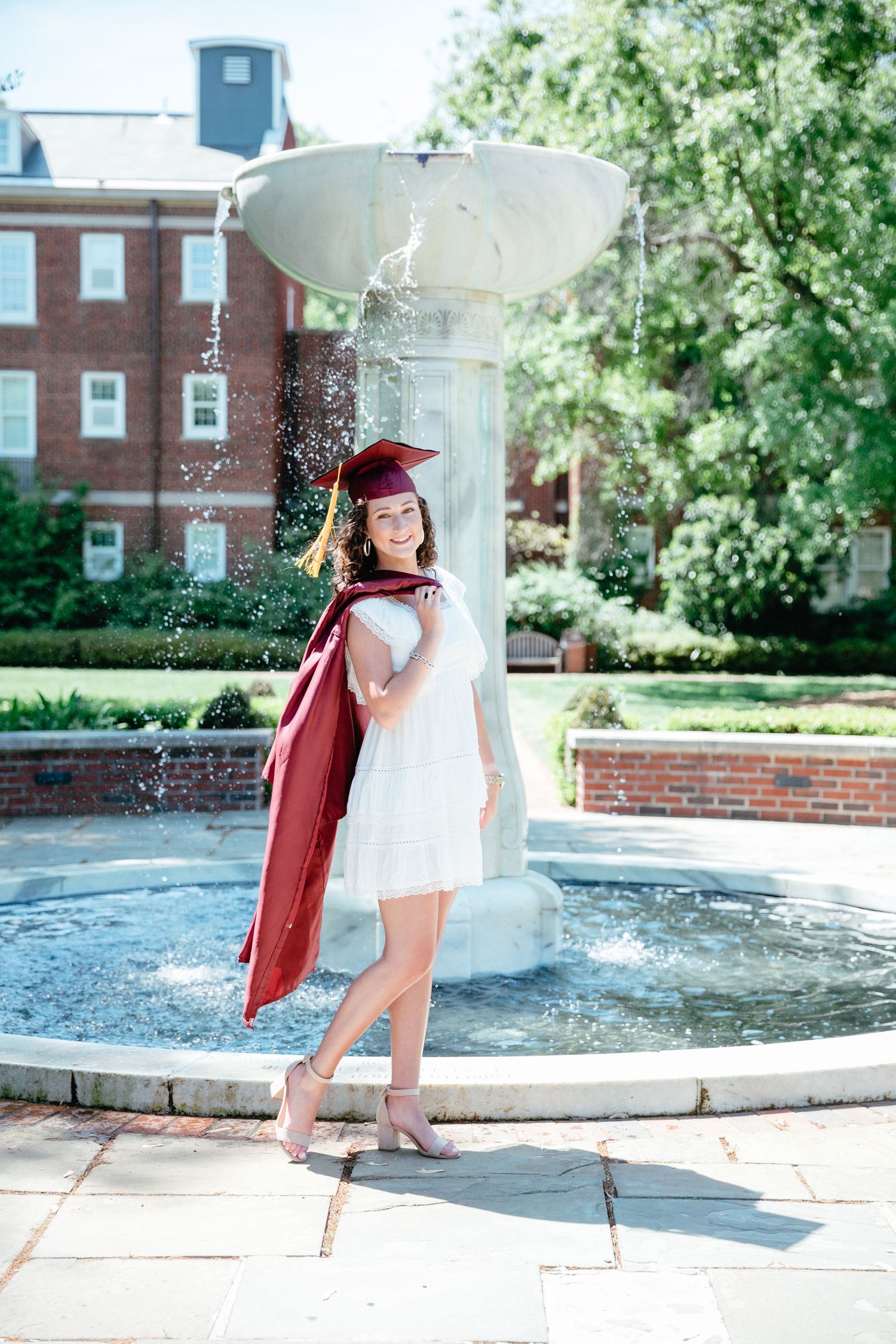 A graduate in cap and gown poses playfully by a fountain on campus.
