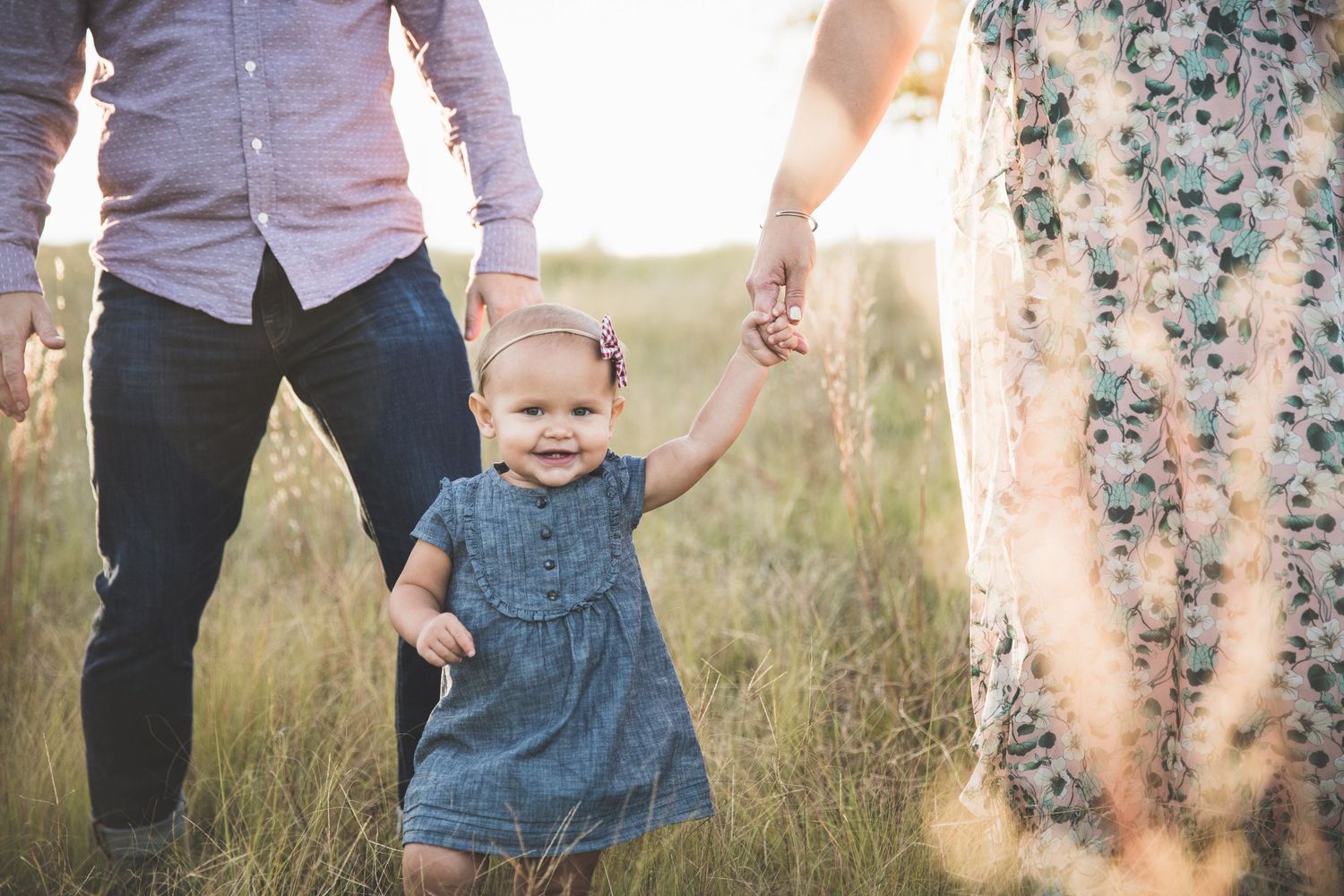 A small child in a denim dress walks through tall grass at sunset.